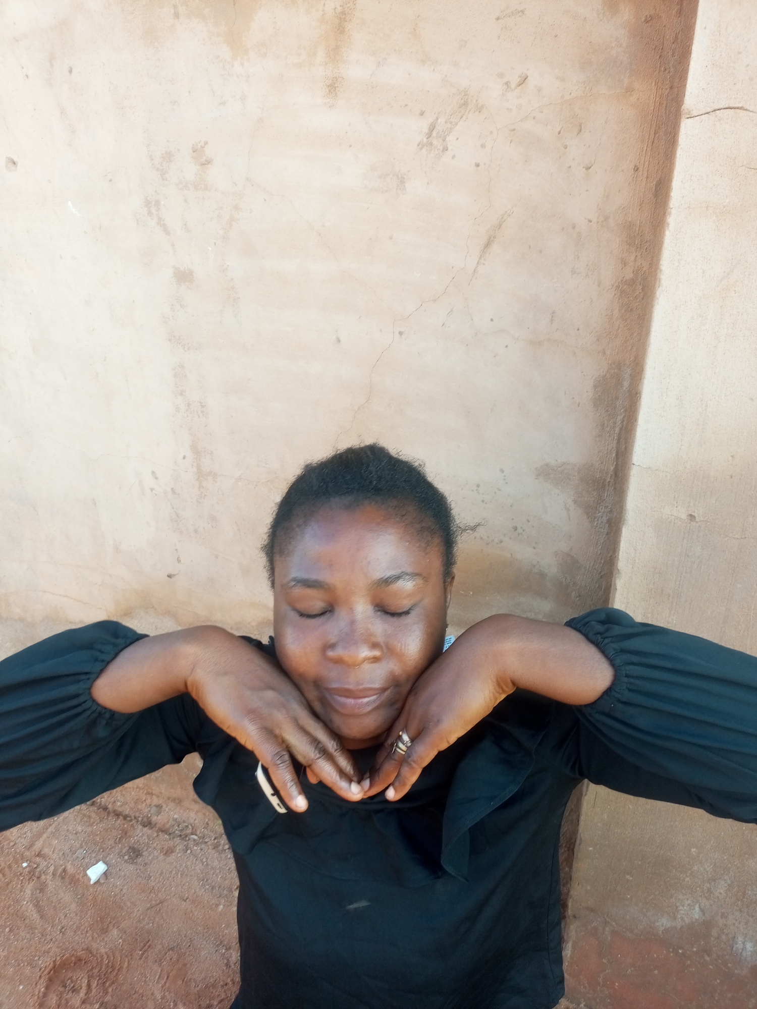 A woman with dark skin and her eyes closed is sitting outdoors, leaning her head on her hands. She is wearing a black long-sleeved shirt. Her hands are placed on her chin and cheeks. A ring is visible on her right ring finger. The background is a textured, light-colored wall and a patch of brown dirt. The image does not contain enough information to determine the specific location in Tempure, Nigeria.