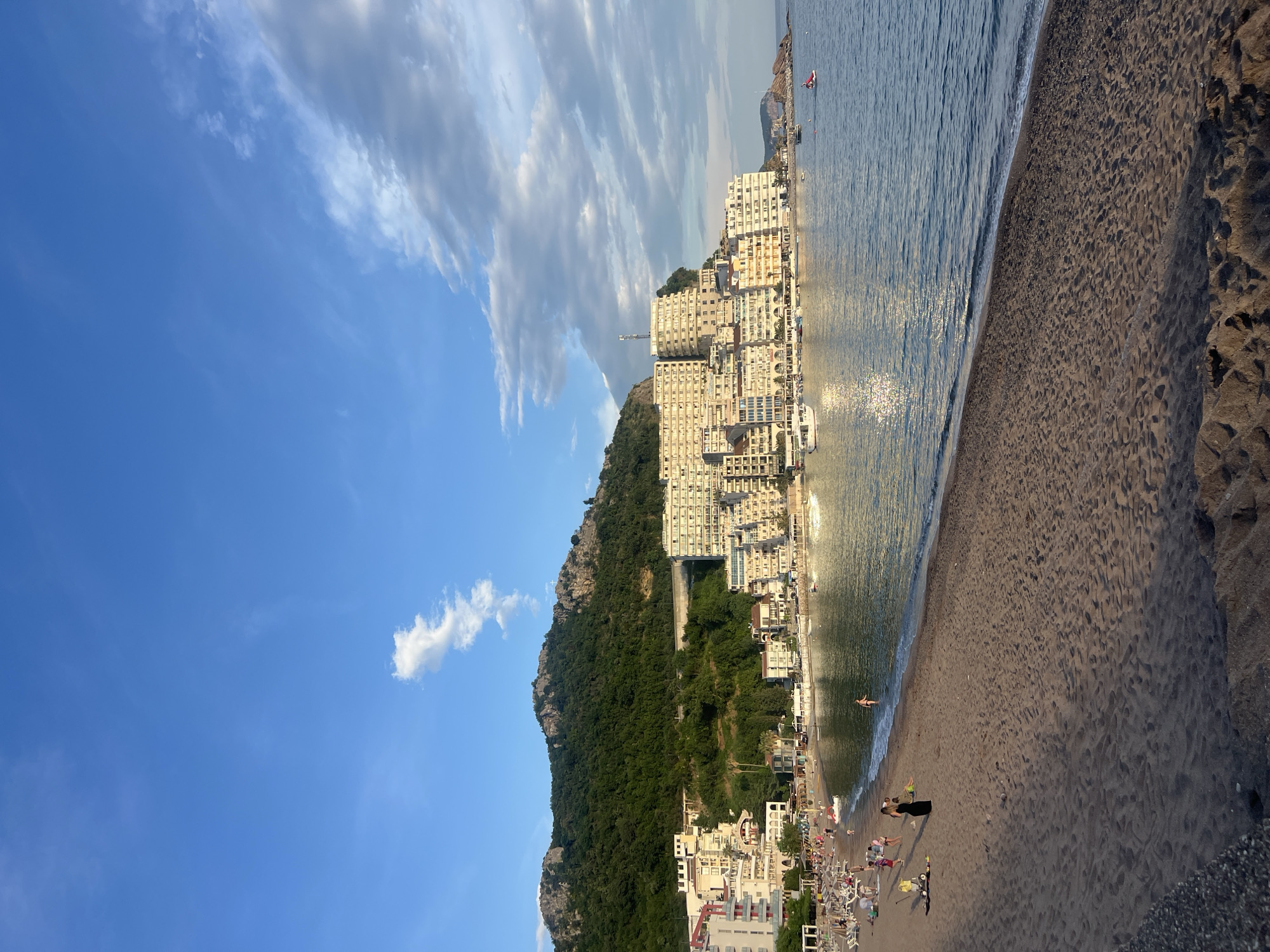 The image shows a beach scene in Rafailovići, Montenegro. The beach is sandy, with visible footprints and patterns in the sand. People are present, some appearing to be sunbathing on beach chairs and others walking or standing near the water's edge. The water is calm, reflecting the sunlight. Adjacent to the beach, a cluster of buildings is built along a hillside, including hotels and apartments. The buildings are of varying heights and architectural styles. A densely forested hill rises behind the buildings. The sky above is blue with scattered white clouds. The image is taken at an angle, as if looking up the coastline.