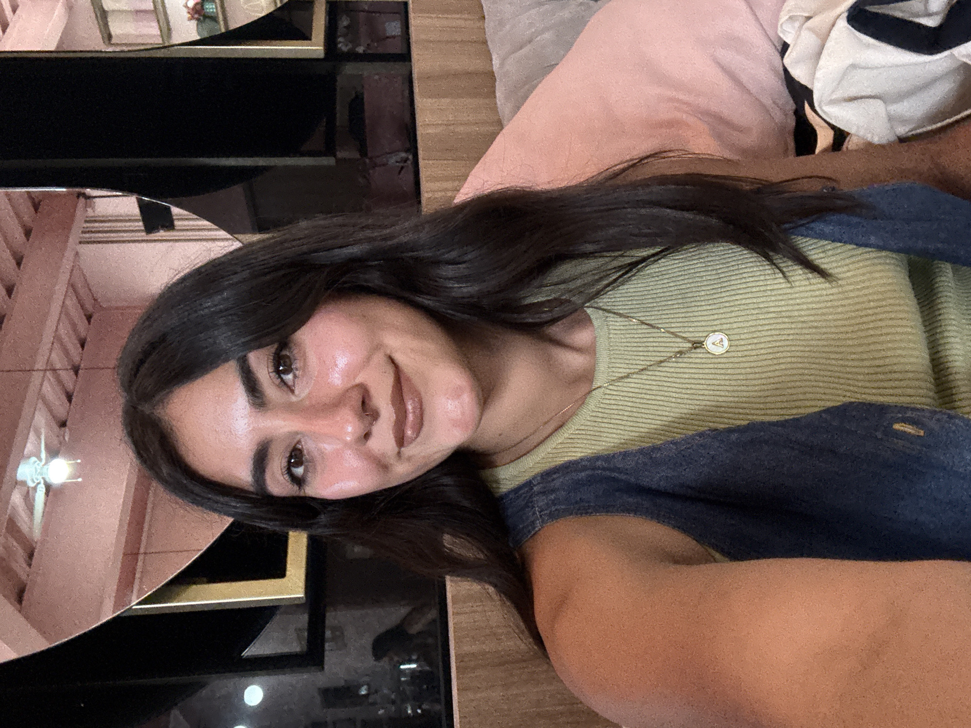 A woman with dark hair and brown eyes is positioned in the foreground, wearing a green striped top and a denim garment. She is smiling at the camera. Behind her, there is a couch with a pink pillow and a striped white-and-black piece of clothing. The background includes a mirror that reflects a building with a pink facade and a ceiling fan. Another part of the mirror reflects a dark area, possibly a room or a window. The location cannot be determined with enough certainty to confirm it is in Puebla, Mexico.