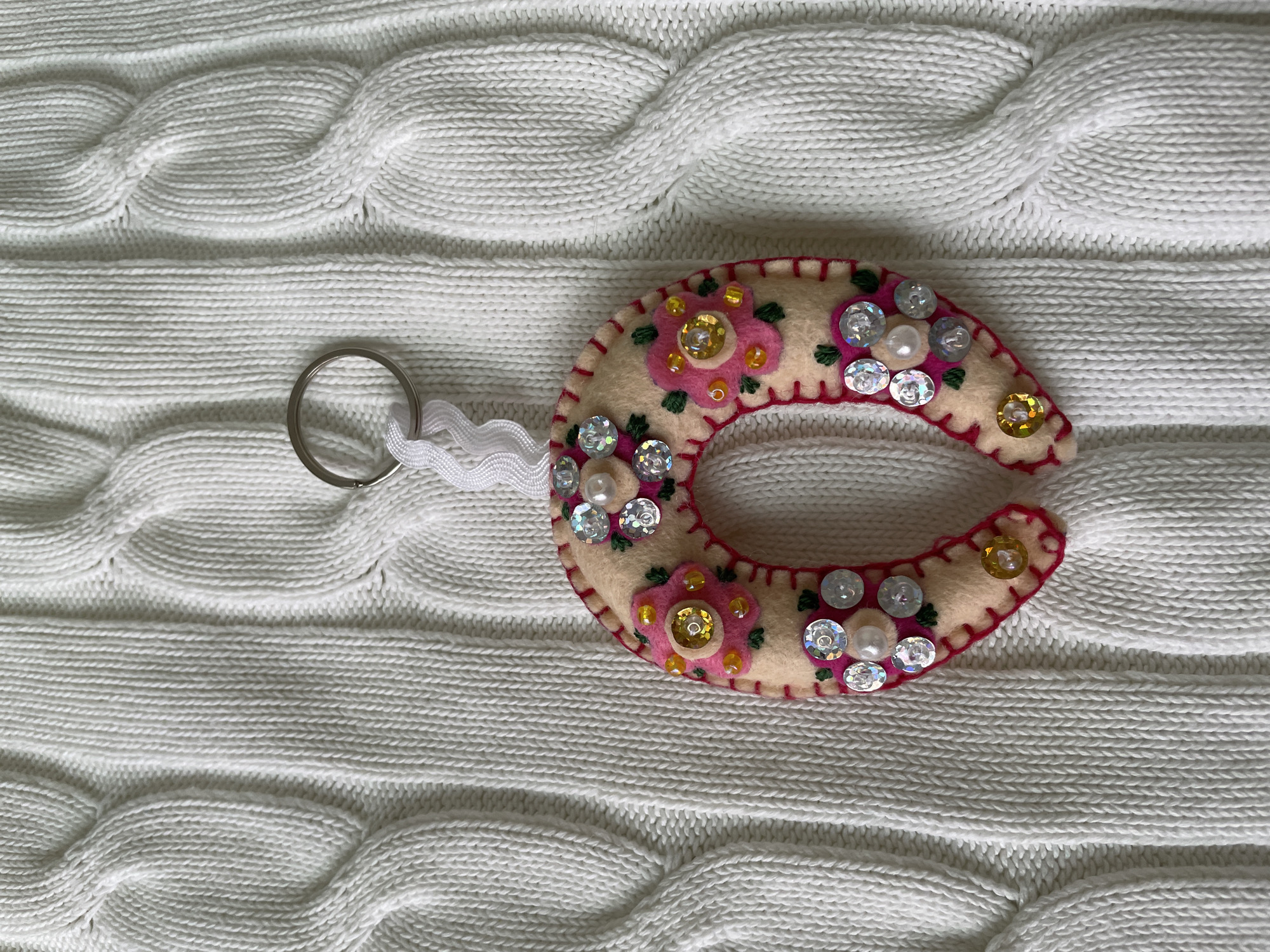 A handmade, horseshoe-shaped keychain lies on a white, cable-knit surface. The keychain is primarily beige felt, decorated with pink embroidered flowers, shiny silver sequins, and small gold beads. A pink embroidered trim borders the edge. The keychain is attached to a silver metal key ring by a short length of white, ribbon-like material. The background is a heavily textured, white cable-knit fabric. The scene is likely indoors in Лесной Пейзаж Кп (шарапово, Russia, based on the image name.