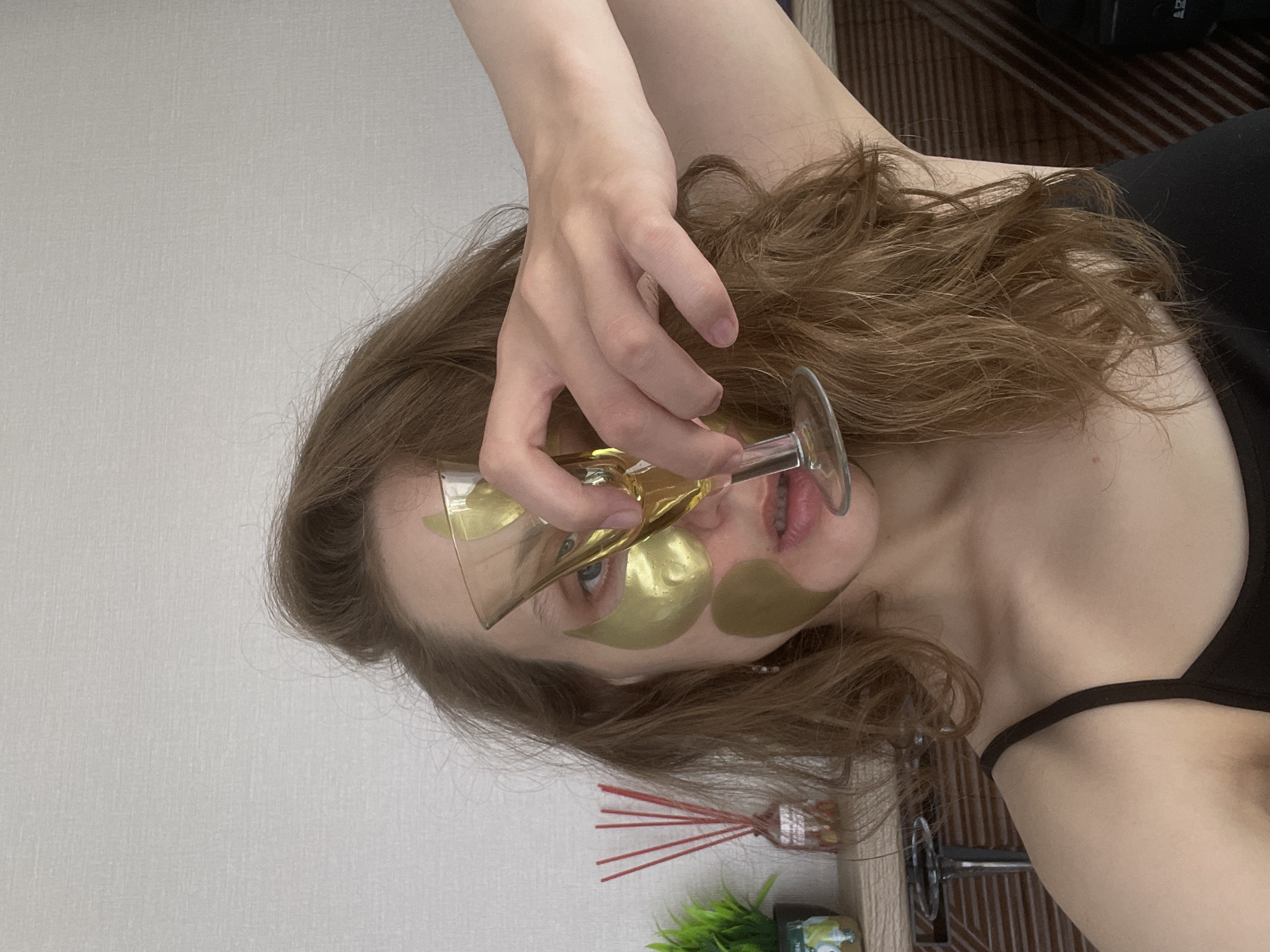 A close-up image shows a young woman in an indoor setting, likely in Подшивалово, Russia. Her head is tilted, with her light brown, wavy hair framing her face and cascading over her shoulder. She has fair skin and visible blue eyes. She is wearing multiple golden, leaf-shaped under-eye patches, suggesting a skincare or relaxation routine.
She holds a clear, stemmed glass, possibly a champagne flute, in her right hand, positioned in front of her face. The base of the glass partially obscures her nose and upper lip, creating a slight distortion of her features. Her mouth is slightly ajar. She wears a black strap top, with a thin strap visible over her right shoulder.
The background features a plain light gray wall to her left and behind her. On a surface to her lower right, a small green potted plant is visible alongside a glass diffuser bottle with several red reeds. Beyond these items, a dark, textured surface, possibly a piece of furniture, and an indistinct dark object are present. The image captures a moment of personal care and domestic relaxation. There is no visible text.
