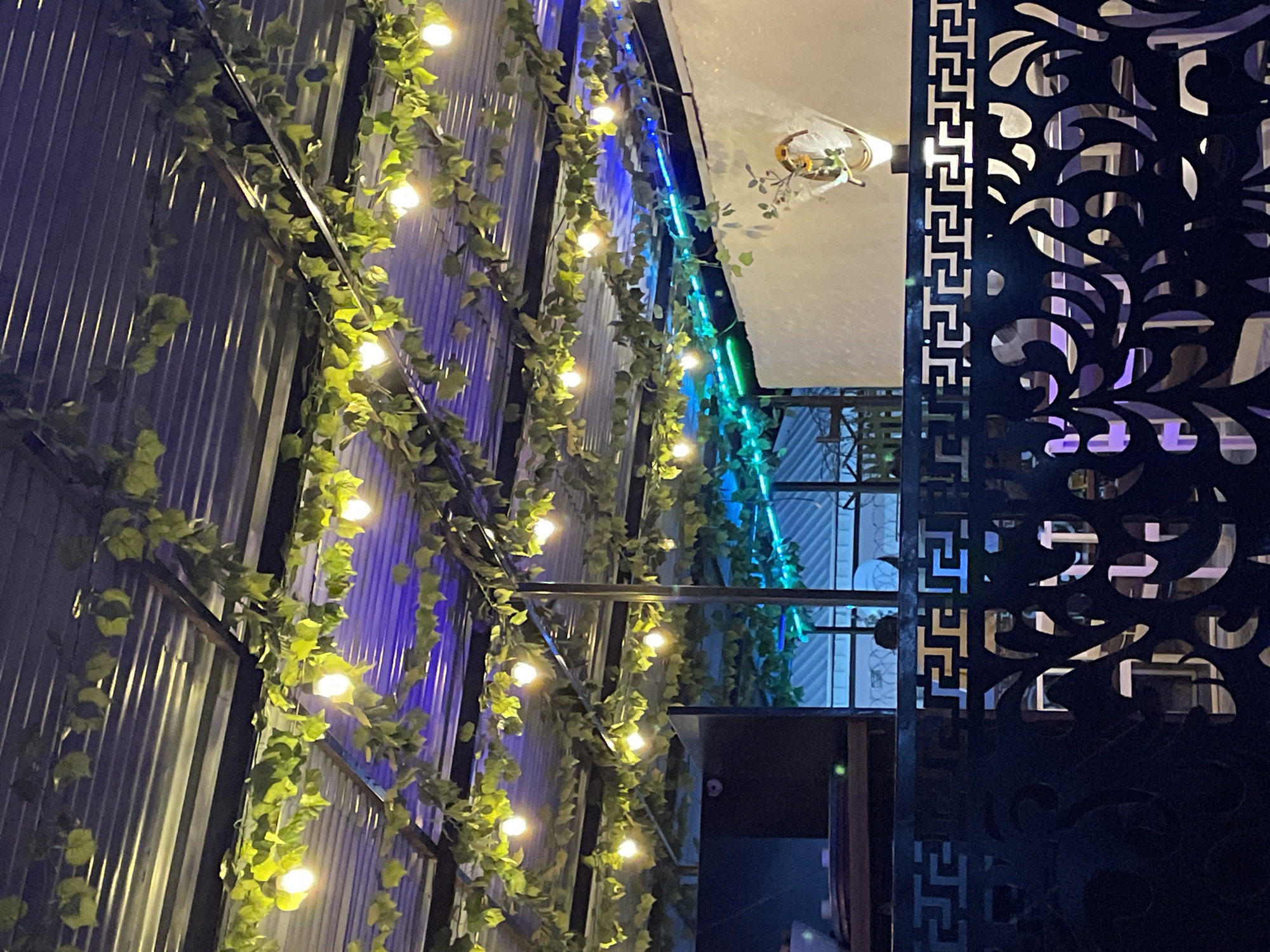 The image shows an outdoor setting, likely a restaurant or bar, located in Jos, Nigeria. A corrugated metal wall on the left is adorned with green vines and small lights, creating a decorative effect. Blue and green lights are strung along the wall, adding to the ambiance. A dark, ornate metal screen or fence is visible on the right, with a complex, flowing pattern. Through the screen, one can see a building with barred windows, suggesting it is enclosed. A partially visible doorway leads into a darker interior space. Overhead, a white canopy or ceiling is visible, with a decorative light fixture hanging down. The overall impression is of a stylish outdoor space with a mix of natural and artificial lighting.