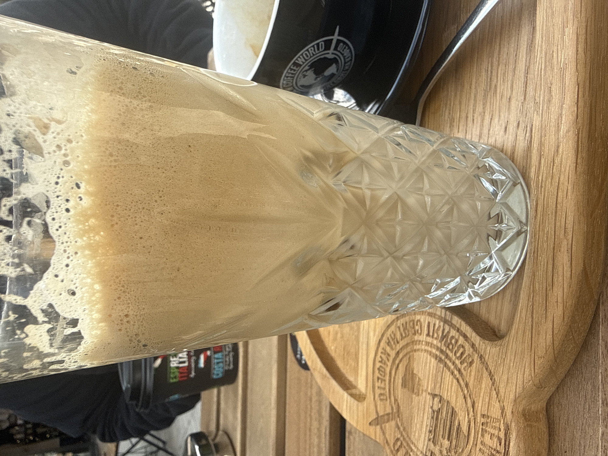 The image shows a glass of iced coffee or similar beverage with a thick foam layer, placed on a wooden serving board that appears to be custom-made with coffee-related text in Bulgarian. The wood grain is clearly visible. Adjacent to the beverage are a black coffee cup with the logo "COFFEE WORLD" and a spoon. Background elements indicate an outdoor setting, possibly a café in Stara Zagora, Bulgaria. A Costa Coffee syrup bottle is visible in the background. A person's arm and clothing are seen in the lower left. The image appears to be taken at eye level.
