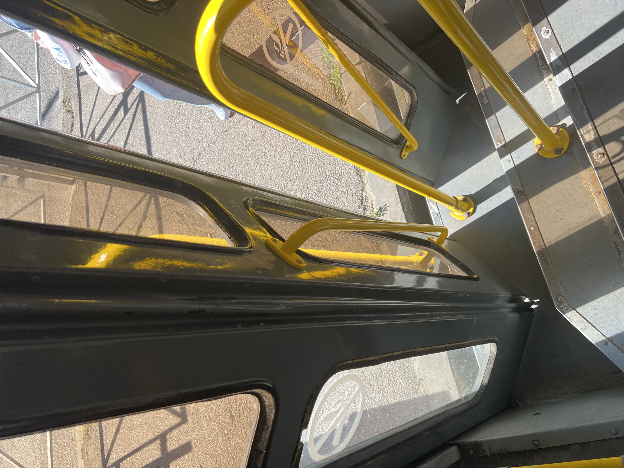 The image shows the interior of a vehicle, likely a bus or tram, with a focus on the doorway and steps. Bright yellow handrails are prominent, guiding passengers up or down a set of metal steps. The steps are worn, showing signs of use. Through the large windows of the door, the exterior environment is visible, including asphalt ground, sunlight casting shadows, and a hint of a person with a light-colored bag. Two circular white stickers with a red diagonal line through a cigarette symbol, indicating "no smoking," are visible on the glass panels of the door. The overall impression is one of public transportation infrastructure.