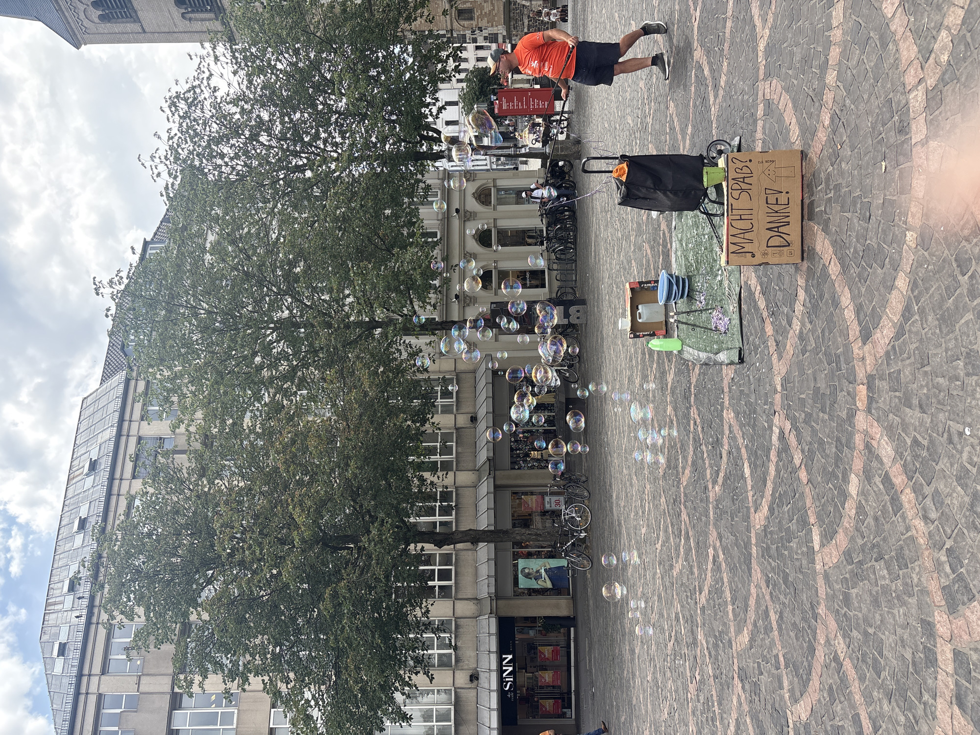 The image depicts a street performer in Bonn, Germany, creating bubbles in an outdoor plaza. The performer, a man wearing an orange t-shirt and dark shorts, is holding a wand and appears to be in motion, possibly mid-action as he blows bubbles. He is on a cobblestone ground, with a large tree and buildings forming the backdrop. Several bubbles of various sizes are visible, catching the light and reflecting colors.
To the right of the performer, there is a cardboard box on the ground with handwritten text. The text, written in German, says "MACHT SPAB? DANKE!". This translates to "Having fun? Thanks!". Next to the box, there is a trolley bag, a jug, and some cleaning supplies, suggesting this is the performer's setup. In the background, there are more buildings, bicycles parked along the street, and other people, indicating a public space likely frequented by pedestrians. The sky is cloudy, suggesting it could be a partly cloudy day. The overall scene evokes a cheerful and playful atmosphere, with the bubbles adding a touch of whimsy to the urban environment. The time of day appears to be daytime, given the lighting conditions.