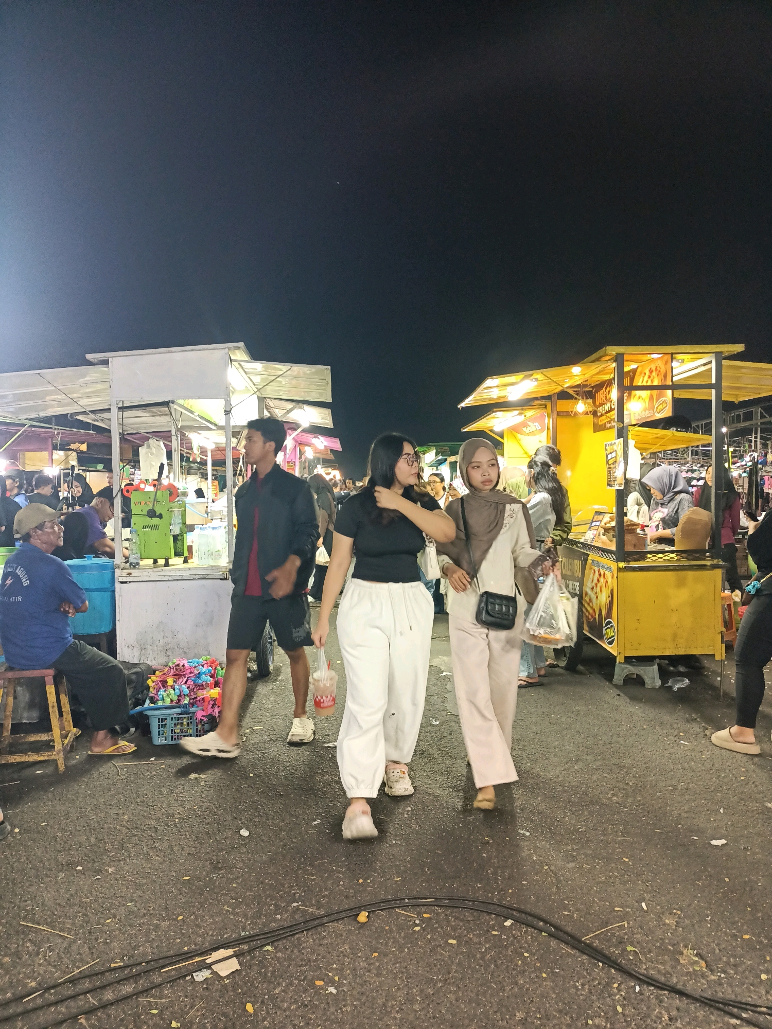 The image captures a bustling night market scene in Surabaya, Indonesia, likely in an open-air location with food stalls. Two young women are the primary focus, walking towards the viewer from the right. The woman on the left wears a black t-shirt and white loose-fitting pants, while the woman on the right is in a beige hijab and a cream-colored outfit, carrying a plastic bag. They are walking on a paved street littered with debris.
In the background, several food stalls are illuminated by bright lights, attracting customers. One stall on the right is yellow and prominently displays images of food. Another stall to the left features a green juicing machine. A man wearing a blue shirt is seated on a stool to the far left, facing away from the camera, with a blue container next to him. A basket filled with colorful toys is visible in the foreground on the left. The overall atmosphere suggests a lively evening market with various vendors and patrons.