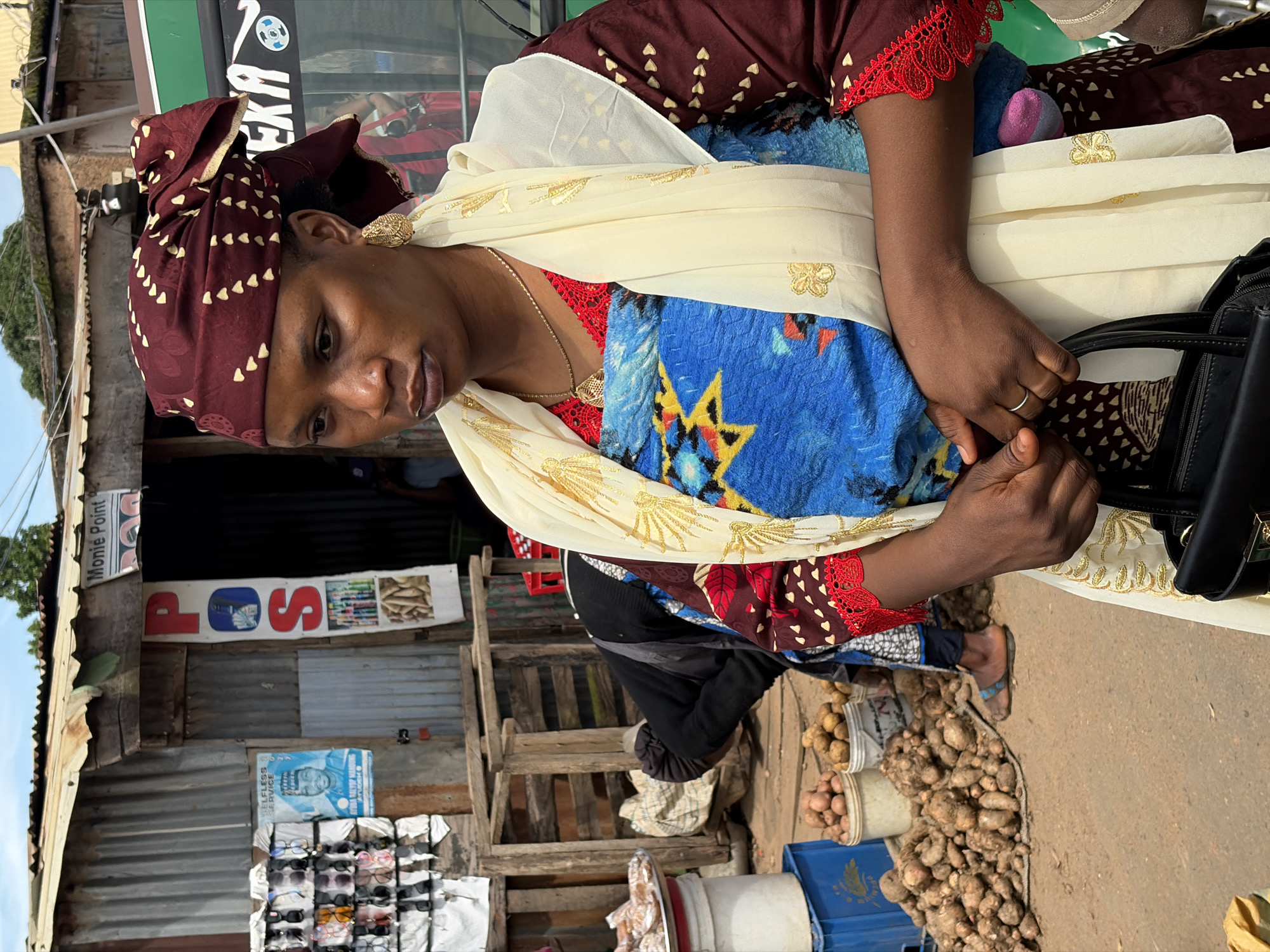 A woman in a maroon and gold headscarf and a red and maroon patterned dress with gold trim stands in front of a market stall in Gura Top, Nigeria. She wears a cream-colored scarf with gold embroidery draped over her shoulders. A blue and yellow patterned cloth is wrapped around her torso. She is holding a black handbag. In the background, there is a stall with various items for sale, including sunglasses, posters, and produce such as potatoes and yams. Other people are visible in the background, as well as a green vehicle. The ground is dirt.