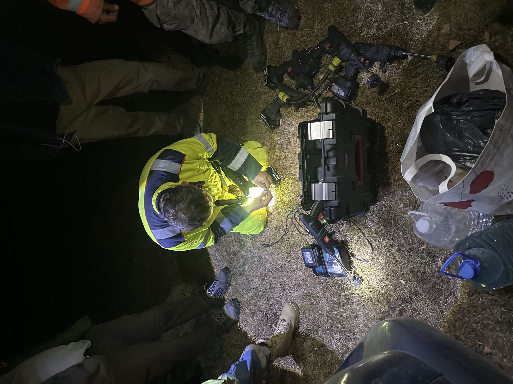 In Bijele Poljane, Montenegro, at night, a group of individuals are gathered around a man wearing a yellow and blue high-visibility jacket. The man is kneeling on dry grass and appears to be working with electronic equipment, holding a flashlight and a small device. Surrounding him on the ground are several power tools, including drills, battery chargers, and a black toolbox. The scene is illuminated by flashlights and a work light, casting strong shadows. Two large plastic water bottles, one with a blue cap and the other clear, are visible in the foreground, along with a white reusable bag containing a black trash bag. The overall environment suggests an outdoor work or repair situation, possibly in a semi-rural or undeveloped area.