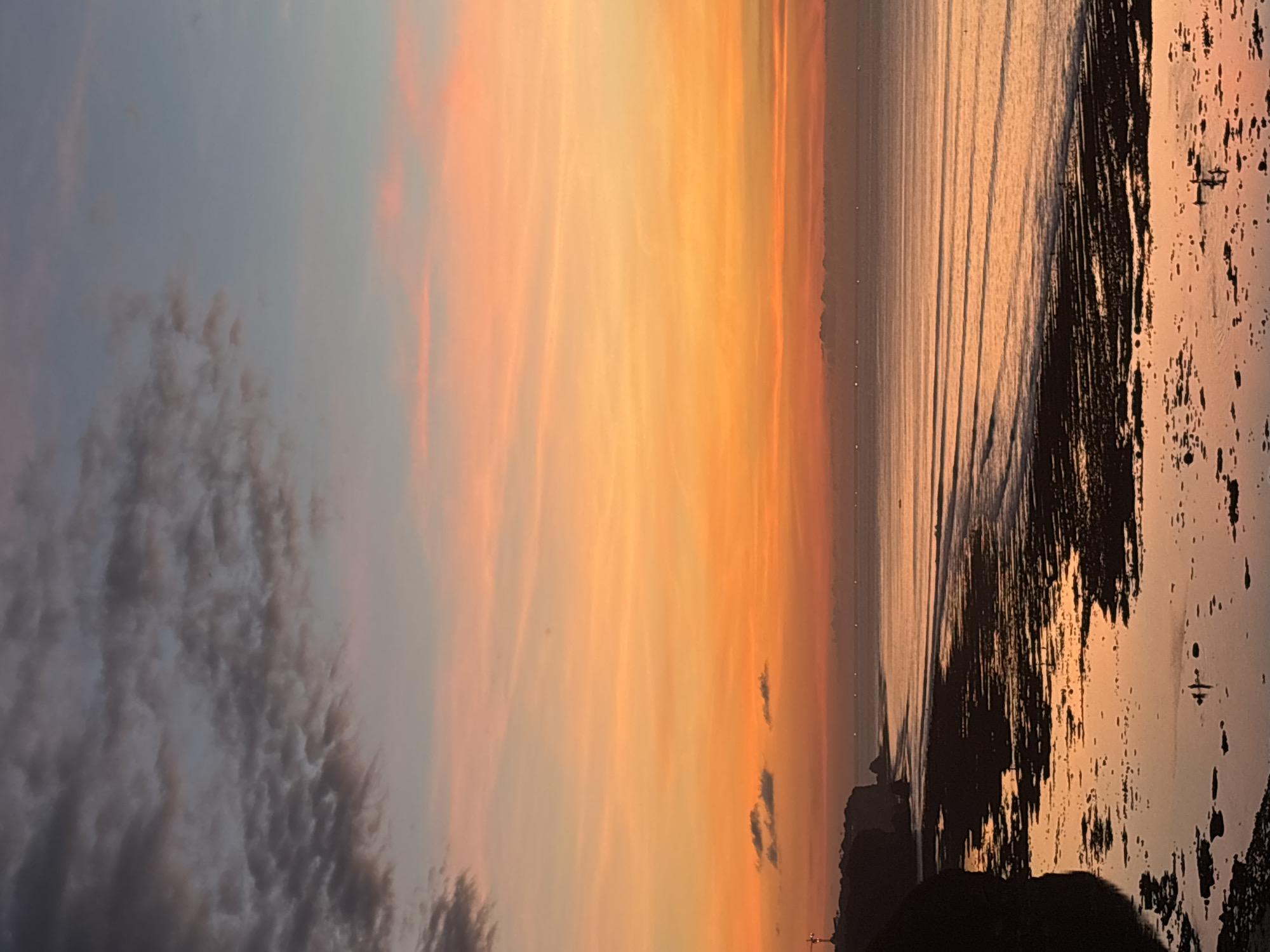 This media file captures a serene coastal landscape during either sunrise or sunset, characterized by a striking color gradient across the sky and its reflection on the water.
The sky, occupying the upper portion of the image, transitions from a soft blue-grey on the left to vibrant oranges, yellows, and peaches towards the right, indicative of a low sun. Scattered clouds are visible, ranging from dark, textured cumulus formations on the upper left to wispy, elongated streaks colored by the sunlight.
Below the horizon, the ocean reflects these warm hues, displaying subtle ripples or wave patterns across its surface. A faint, horizontal line of small, dark objects, possibly distant boats or buoys, dots the water near the horizon line.
The bottom right section of the image features a dark, rugged shoreline or beach, appearing to be wet and reflecting the light, with irregular dark shapes that could be rocks or damp sand. Along the horizon, a prominent silhouetted landmass or cliff is visible, on top of which stands a distinct cross-like structure, strongly suggesting a temple or monument, possibly the Uluwatu Temple given the Kuta Selatan, Indonesia context.
There are no discernible people or visible text in the image. The overall mood is tranquil and contemplative, highlighting the natural beauty of the coastal scene at dusk or dawn.