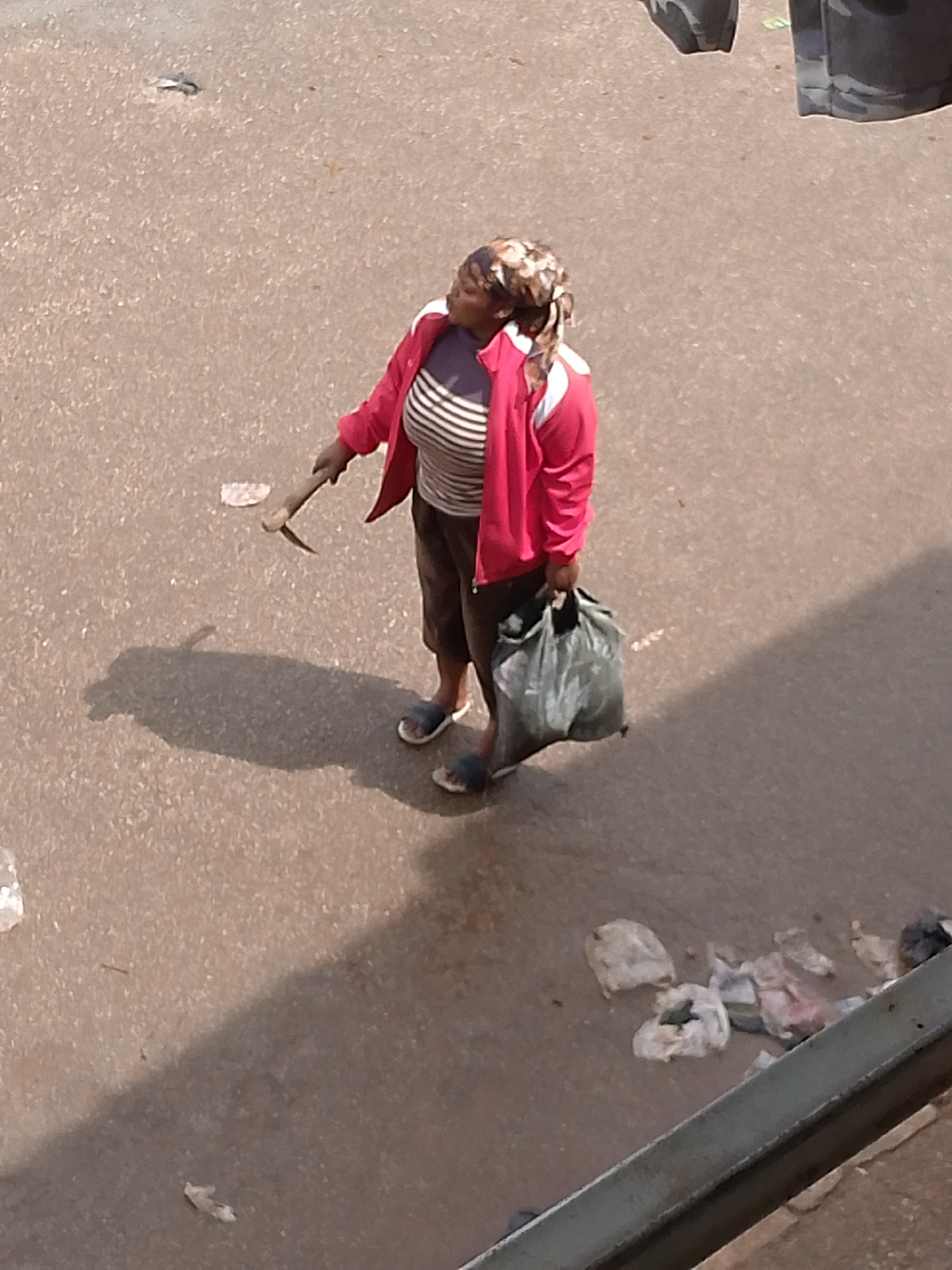 An overhead view captures a woman standing on a paved surface, likely a street or pathway, in Bukuru, Nigeria. She is wearing a pink jacket over a striped shirt and dark trousers. On her left hand, she holds a pickaxe. In her right hand, she carries a large, dark garbage bag. The ground around her is littered with various pieces of refuse. A prominent shadow is cast by the woman and her tool onto the ground. The lighting suggests it is daytime, with the sun casting strong shadows. The overall scene depicts an urban environment with evidence of littering.