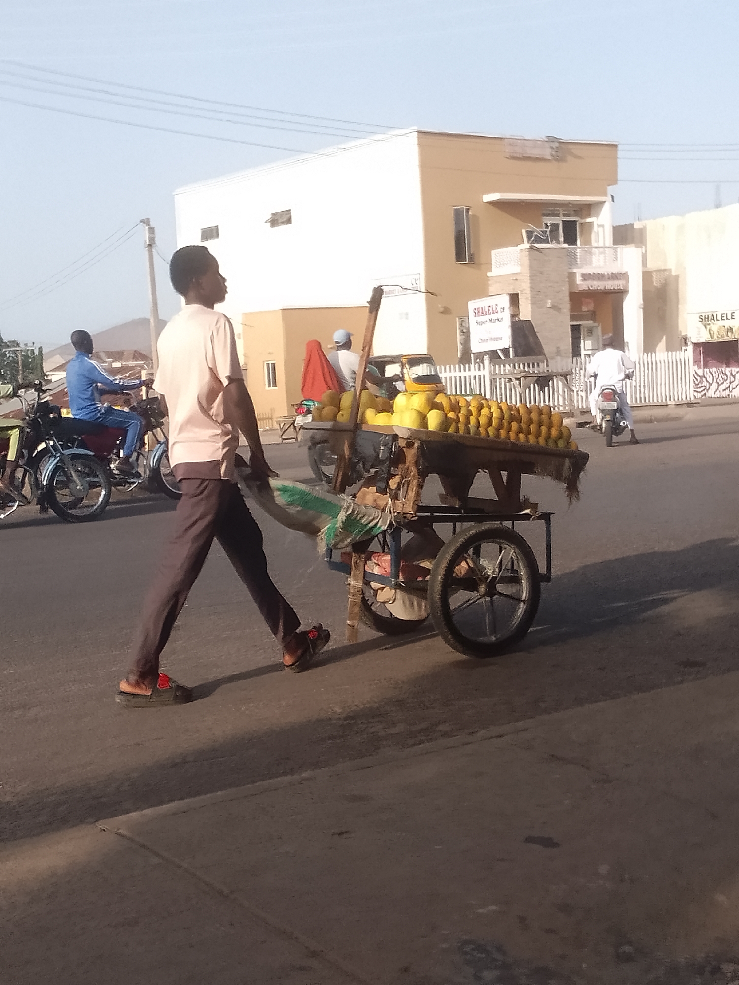 A man pulls a fruit cart along a city street. The cart is piled high with yellow fruits. He wears a light pink shirt, dark pants, and sandals. In the background are buildings, motorcycles, and other people on motorcycles. Signs reading "SHALELE" are visible. The location is Pantame, Nigeria.