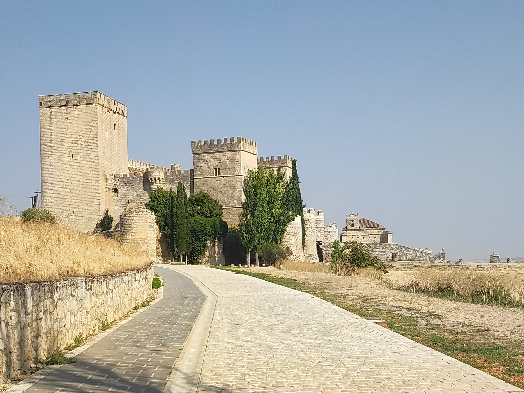 The image shows the Castle of Ampudia, located in the town of Ampudia, Palencia, Spain. The castle is a well-preserved medieval fortress made of stone, with a prominent square tower and crenellated walls. A paved pathway leads towards the castle entrance. The surrounding landscape is dry and arid, with sparse vegetation and a clear blue sky. There are no people or animals visible in the image. The overall impression is one of historical significance and architectural grandeur.