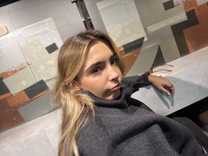 The image depicts a female individual in an indoor setting in Vaulx-en-Velin, France. The subject, positioned centrally, has long, light brown hair with blonde highlights, styled straight. She is wearing a dark grey, high-neck, long-sleeved garment of a textured fabric. Her right arm is bent, with her hand resting on a light grey, solid surface, possibly a bench or counter. A dark-colored ring is visible on her right ring finger, and a multi-strand light brown bracelet is on her right wrist. Her fingernails are painted a light color. She looks directly at the camera with a neutral expression, her lips slightly pursed. The background consists of a wall with an abstract geometric design featuring shapes in shades of grey, beige, orange, and dark grey. An upright dark structural element is visible on the far right. The lighting suggests an artificial indoor environment.