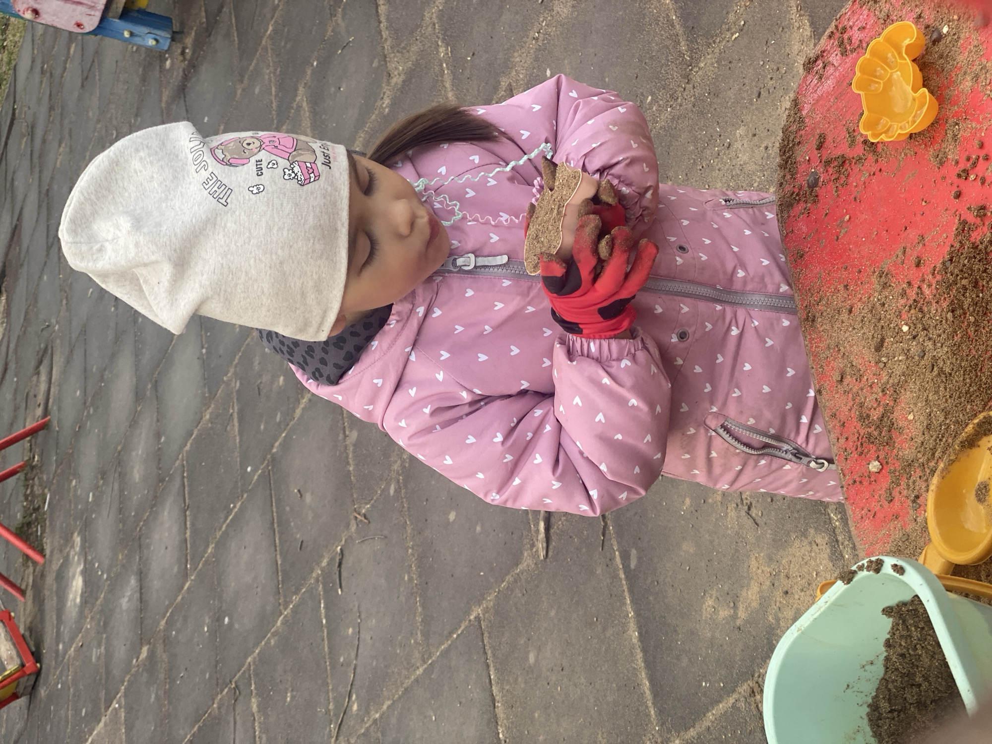 This outdoor, eye-level shot in Chaikovsky, Russia, captures a young child intently engaged in sand play. The child, likely a girl, is dressed in warm attire, suggesting cool weather. She wears a light gray beanie featuring a cartoon bear with headphones and partially visible text that reads "THE JOU," "CUTE," and "Just En." Her pink puffer jacket is adorned with small white heart patterns, and she has red and black gloves on her hands.
The child's gaze is directed downwards, and her eyes appear closed or focused on the activity. She holds a broken piece of what looks like a molded sand creation in her gloved hands, which are covered in sand. In front of her is a red surface, possibly a small table or sandbox edge, also heavily covered in sand. An orange plastic sand mold, shaped like an animal, rests on this red surface.
To the bottom right, a light blue plastic bucket filled with sand and a yellow plastic spade or scoop handle are visible, further indicating a sand play area. The ground beneath the child is a dark gray paved surface composed of rectangular blocks. A turquoise wavy ribbon or string is draped near the child's chest. The overall scene conveys a moment of focused, imaginative play.