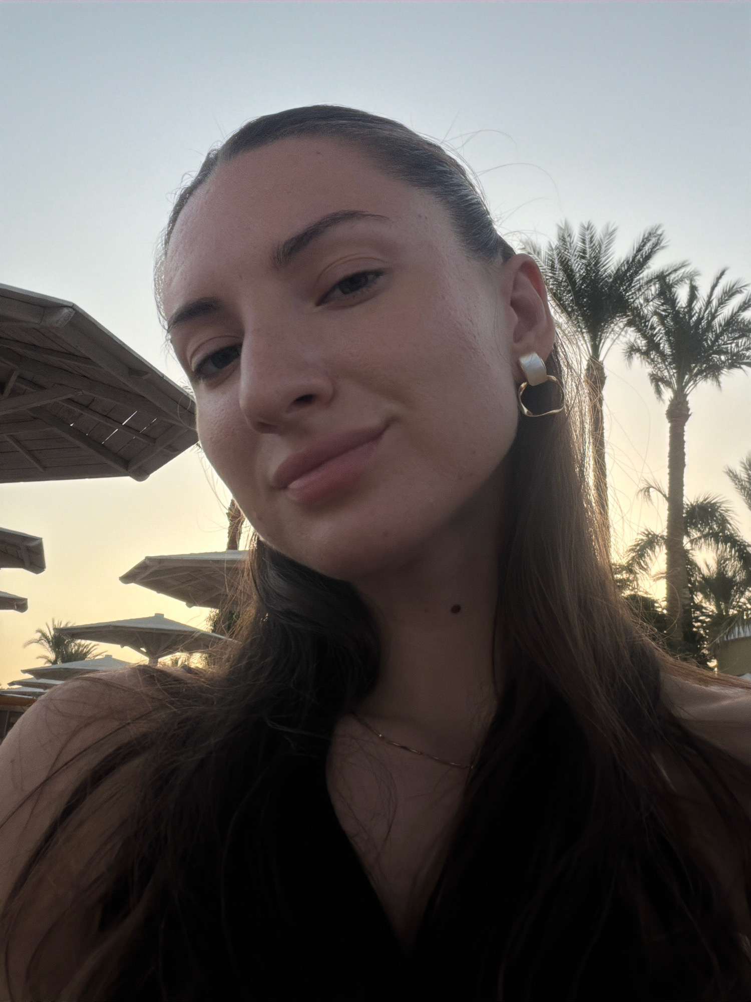 A young woman with long, dark brown hair is taking a selfie outdoors at dusk. She is wearing a dark top and a gold earring. Behind her, palm trees and beach umbrellas are visible, suggesting a resort setting. The sky is a hazy orange, characteristic of sunset or sunrise. The image appears to have been taken in Sharm El Sheikh, Egypt, given the environmental cues.