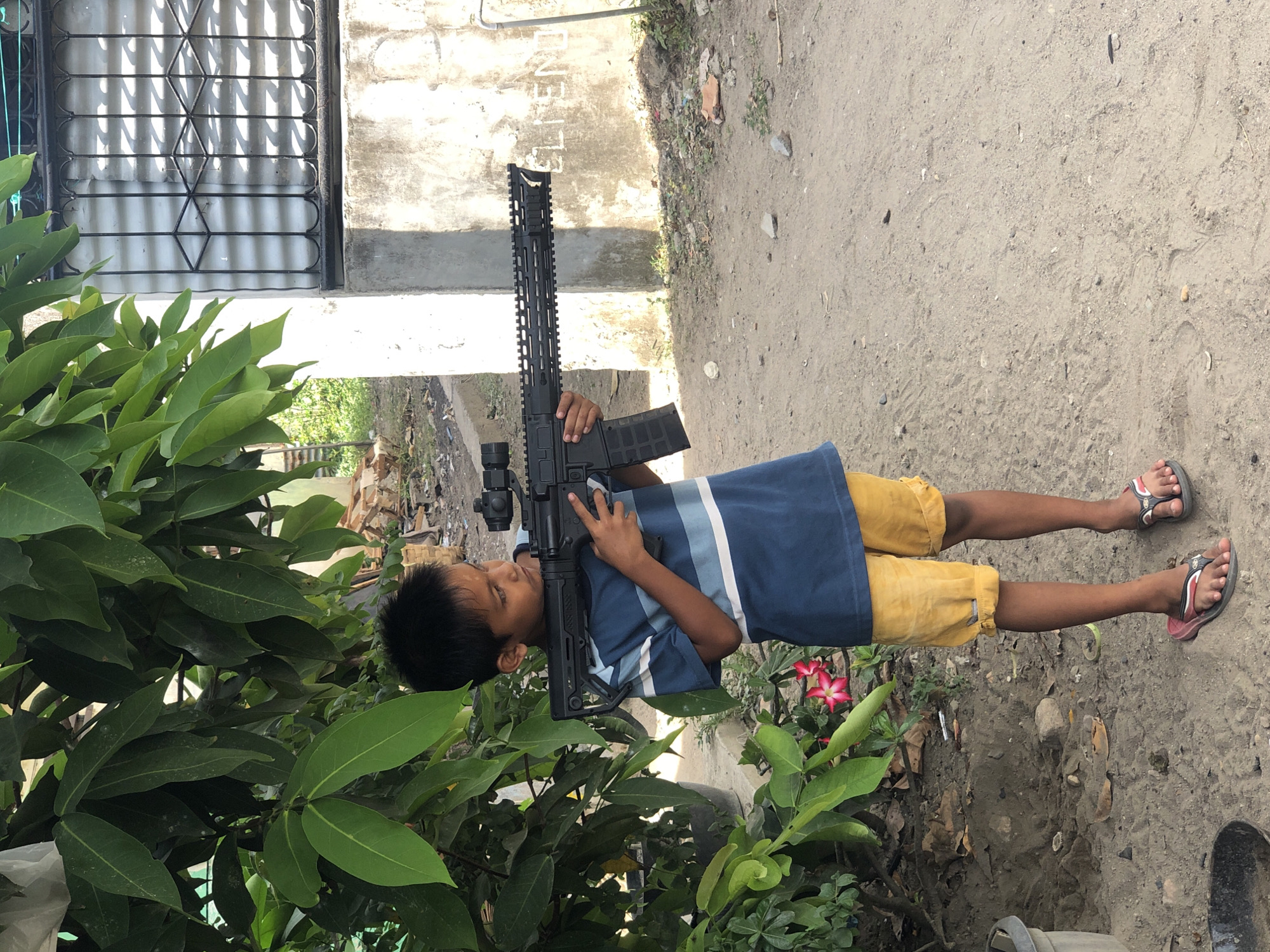A young boy stands outdoors, holding a black rifle. He wears a blue and white striped shirt, yellow shorts, and sandals. He is positioned near a building with a window covered by a metal grille, and adjacent to vegetation. The ground appears to be dirt and gravel. The location is likely Indonesia, but not specified within the picture.