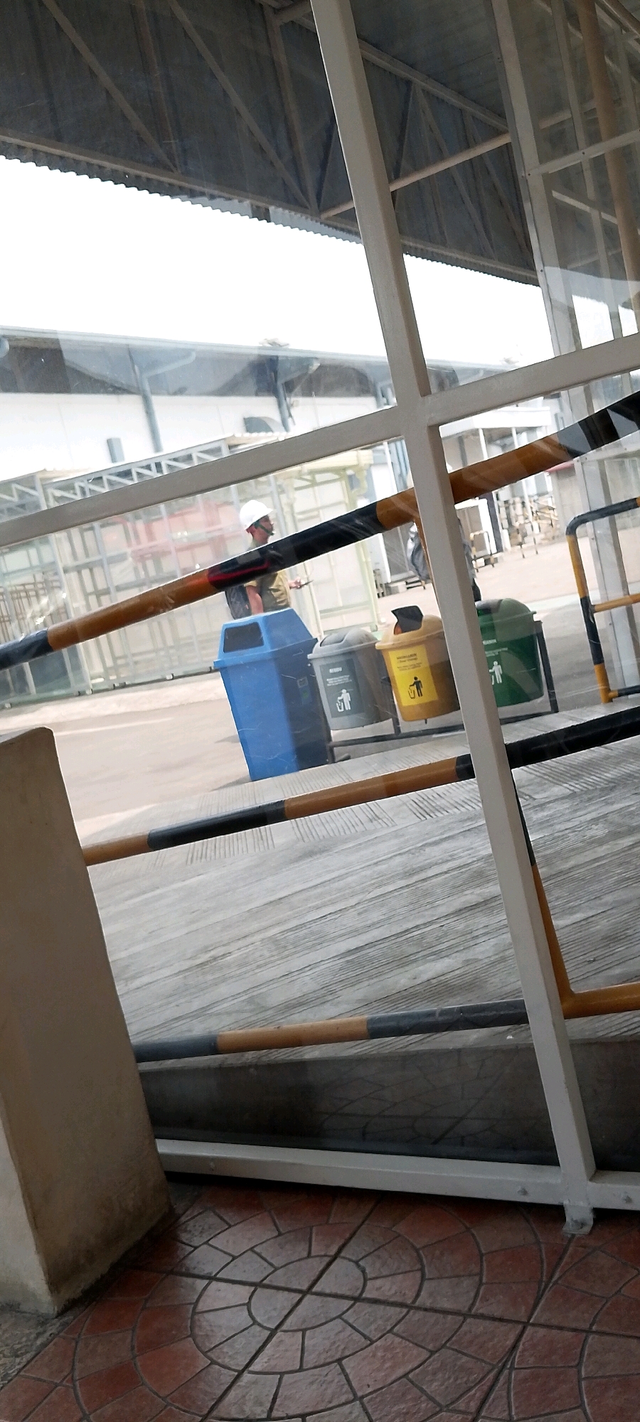 The image appears to be taken in an outdoor area with a covered structure above, possibly a walkway or a waiting area. The scene is viewed through a glass partition with white framing.
In the background, a person wearing a white hard hat and a dark shirt is walking. They are holding a mobile device in their right hand. In front of them, there are several colored trash bins: a blue bin, a gray bin with a recycling symbol, a yellow bin with a recycling symbol and a graphic of a person throwing trash, and a green bin with a recycling symbol and a graphic of a person throwing trash.
The ground is paved with concrete, and in the foreground, there is a tiled area with a circular pattern. Several horizontal bars, some wrapped in black and yellow tape, are present, possibly acting as barriers or railings.
The overall impression is of a public space, likely within a facility or transportation hub in Depok, Indonesia, given the context. The lighting suggests it is daytime, with bright light coming from the covered area.