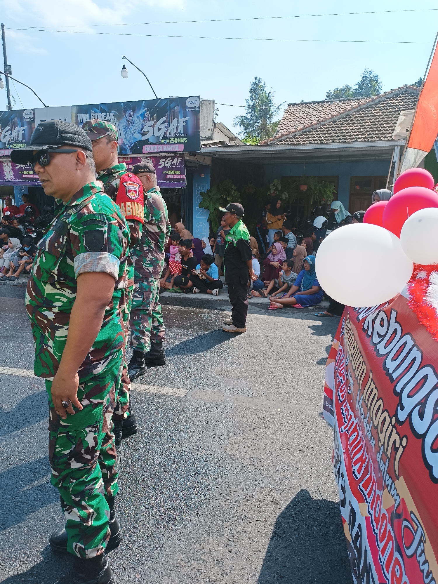 In Imogiri, Indonesia, on a sunny day, a group of people are gathered on the side of a street. In the foreground, two individuals in camouflage military uniforms stand facing left, with one wearing sunglasses and a black cap. Behind them, another person in a similar uniform is visible, with red armbands. A man in a green and black uniform stands to the right of the military personnel, facing forward. A crowd of spectators, including women in hijabs and children, are seated on the roadside. A red and white banner with text is prominently displayed on the right side of the image, and red and white balloons are attached to it. In the background, a building with a tiled roof and blue walls is visible, along with trees and a clear blue sky. A large billboard advertising "WIN STREAK 5GAF ABSOLUTE FAST UP TO 250 MBPS" is also present in the background, indicating a promotional event or local gathering.