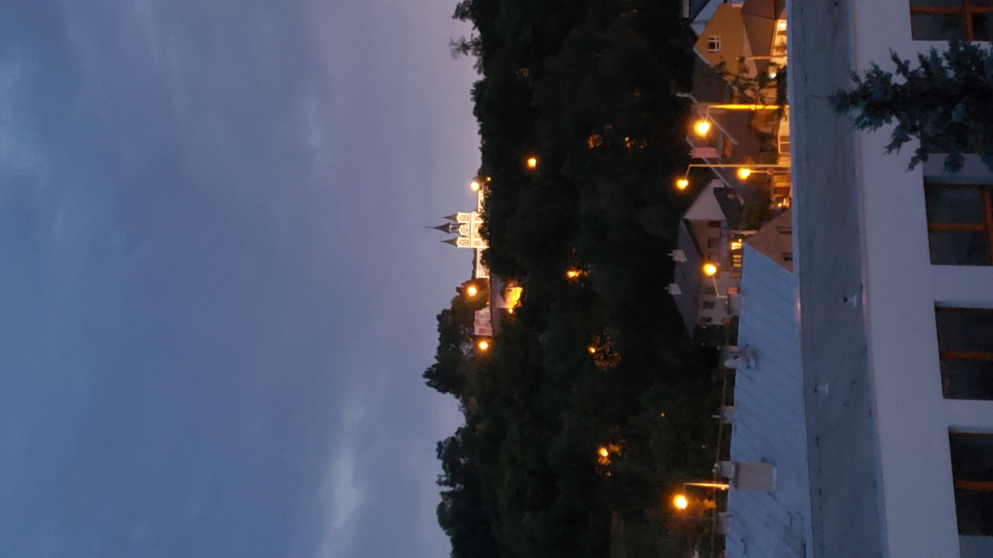 The image captures a twilight view of a city, possibly Prešov, Slovakia, from an elevated perspective. In the foreground, a modern building with large windows is visible. Behind it, a series of rooftops with a light blue metal roof and chimneys can be seen, with several houses nestled behind them. Streetlights with bright yellow bulbs illuminate the scene.
Dominating the middle ground is a dense, dark green hillside covered in trees. At the crest of the hill, a distinctive white building with red accents and multiple spires stands out, illuminated by artificial light. Several other illuminated structures or lights are scattered among the trees on the hillside, suggesting residential areas or other buildings. The sky above is a deep blue, with some subtle cloud formations, indicating the transition from day to night. The overall impression is one of a quiet, European town at dusk.