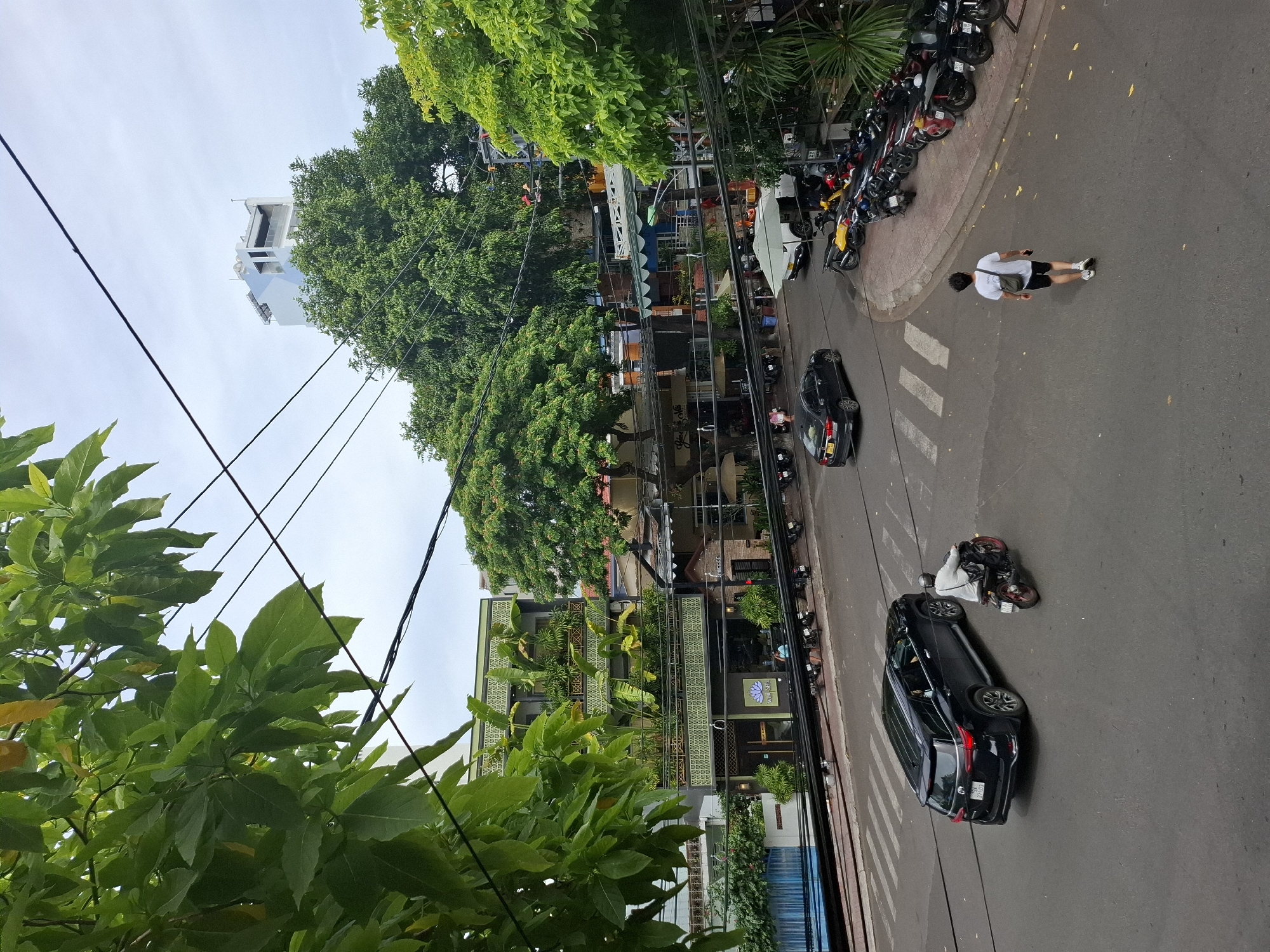 The image shows a street scene in Nha Trang, Vietnam. In the foreground, a black SUV is driving on the left side of the road, followed by a motorcycle with a rider wearing a white shirt and black pants. A pedestrian in a white t-shirt and black shorts is walking away from the camera on the right side of the road. There are several parked motorcycles along the sidewalk. In the background, there are buildings with lush greenery, including trees and plants on balconies. A tall, modern building is visible in the distance. Power lines crisscross the sky. The overall impression is of a busy urban environment with a strong presence of nature.
