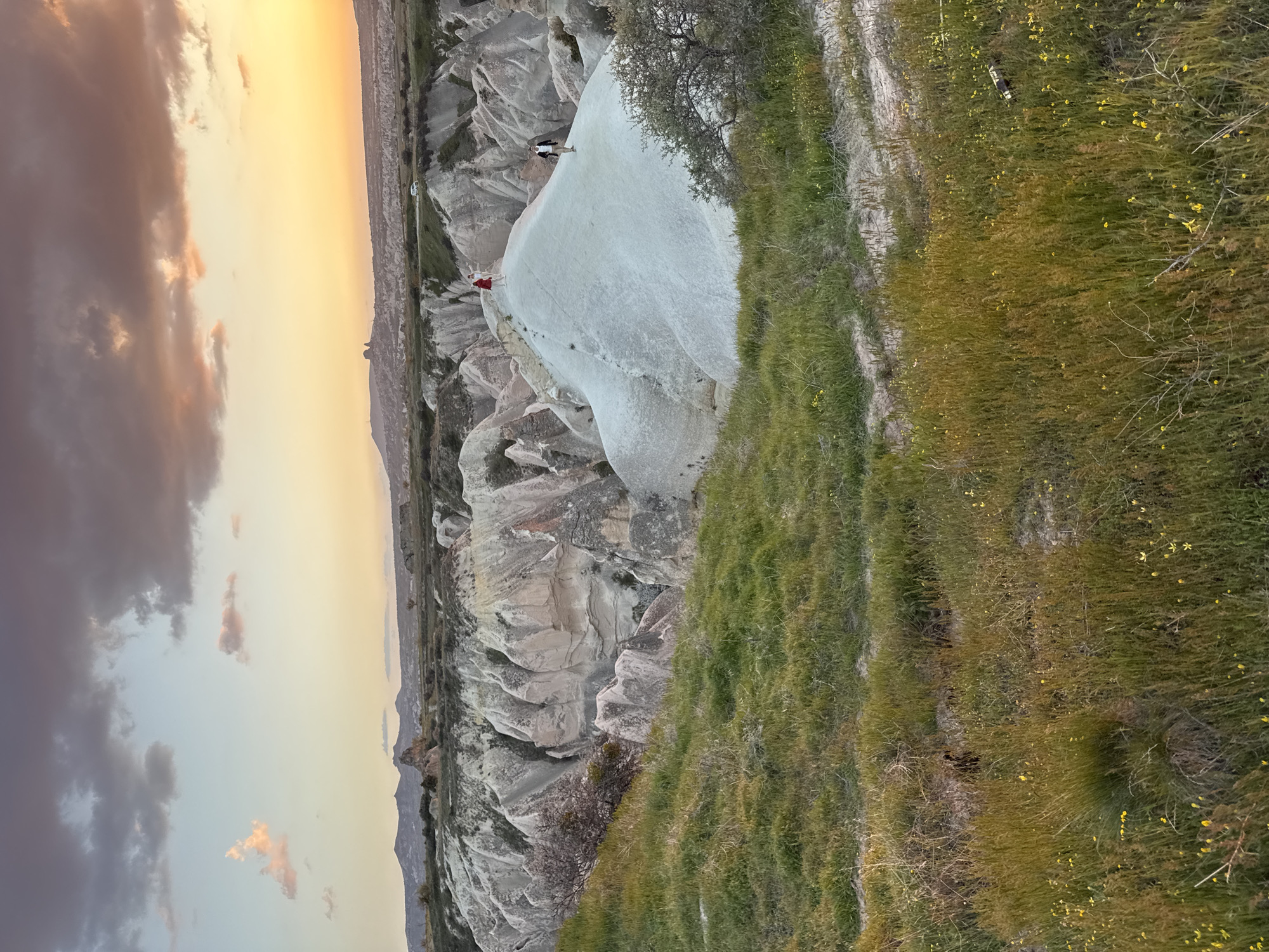 This media file captures a scenic landscape in Ürgüp, Turkey, at either sunrise or sunset, indicated by the warm, golden light illuminating the sky and terrain. The image is composed of unique geological formations, lush foreground, and a vibrant sky.
In the mid-ground, a prominent, large, smooth white rock formation dominates the scene, characterized by its undulating surface. Two individuals are visible on this formation: one dressed in red stands near the edge, looking out over the landscape, while another person in lighter attire is further back. Surrounding this central formation are other irregularly shaped, lighter-colored rock structures typical of the Cappadocia region.
The foreground on the right side of the image is covered with green grass interspersed with small yellow wildflowers, suggesting a verdant slope. A few sparse trees or bushes are also present in this area. In the distance, on the far left of the image (behind the rock formations), the landscape continues with more rolling hills and valleys. A light-colored vehicle, possibly white, can be seen on a distant track or road.
The sky occupies the upper portion of the image, displaying a beautiful gradient of colors. Closer to the horizon, it transitions from a bright orange-yellow to softer peaches and pinks, blending into cooler purplish-gray tones higher up, with scattered clouds adding texture. The overall impression is one of serene natural beauty and vastness, with the people serving as small scale indicators within the grand landscape. No visible text is present.