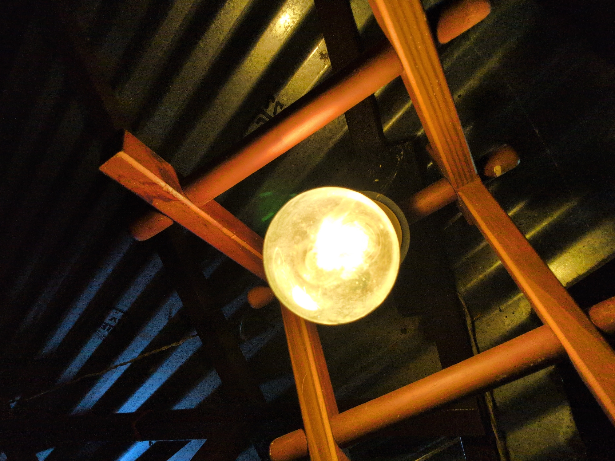 An upward angle shot captures a lit incandescent light bulb suspended from a wooden structure against a dark, corrugated ceiling in Bhagsu, India. The light bulb, emitting a warm yellow glow, is positioned centrally in the frame. Wooden beams, painted a reddish-brown, form a crisscross pattern above, supporting the bulb. The ceiling, likely made of corrugated metal, is dark and textured, with diagonal shadows cast by the wooden structure. The overall impression is one of a rustic interior, possibly a restaurant or a dwelling, with the light bulb providing the primary illumination. No people or animals are visible in the scene.