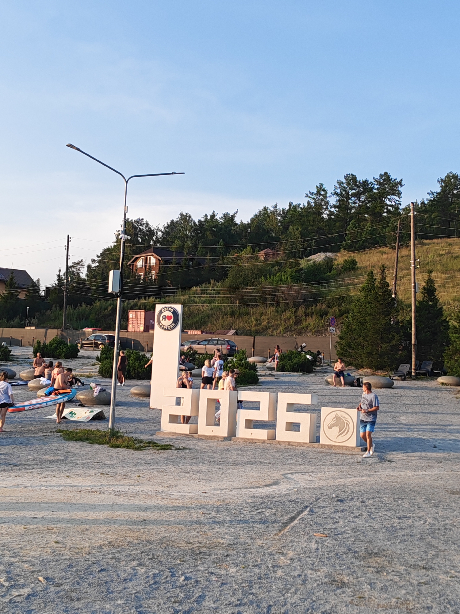 The image depicts a scene at a lakeside resort in Turgoyak, Russia, on a clear day with a bright blue sky. In the foreground, a large, white, three-dimensional sign reads "2026" with a circular emblem featuring a horse's head and the text "Я ЛЮБЛЮ ТУРГОЯК" (I Love Turgoyak) to the left of the number six. The sign is situated on a sandy or gravelly ground surface.
Scattered around the sign are various people, most of whom appear to be vacationers. Several individuals are standing, sitting, or walking. To the left of the sign, a person is holding a paddleboard. Other people are seen on or around large, smooth, grey rocks. In the background, there is a dense hillside covered with trees and bushes. Several houses are visible nestled among the trees on the slope. Power lines and utility poles are also present.
Further in the background, a few vehicles, including cars and a motorcycle, are parked. The overall environment suggests a recreational area, likely a beach or park setting near a body of water, given the presence of the paddleboard and the resort-like atmosphere. The lighting indicates it is likely late afternoon or early evening.