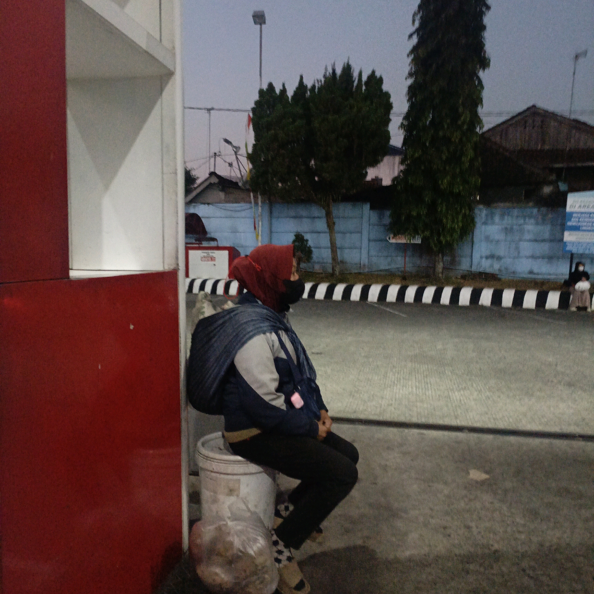 A person wearing a red hijab and a dark jacket sits on a white bucket next to a red and white structure, possibly a gas station in Salatiga, Indonesia. The individual is looking away from the camera with a blue bag slung over their shoulder. A plastic bag filled with items sits on the ground near the bucket. In the background, a blue wall with black and white stripes runs along a paved area. Trees and buildings are visible behind the wall, and a street light stands tall. The sky is overcast, suggesting it might be dusk or early morning.