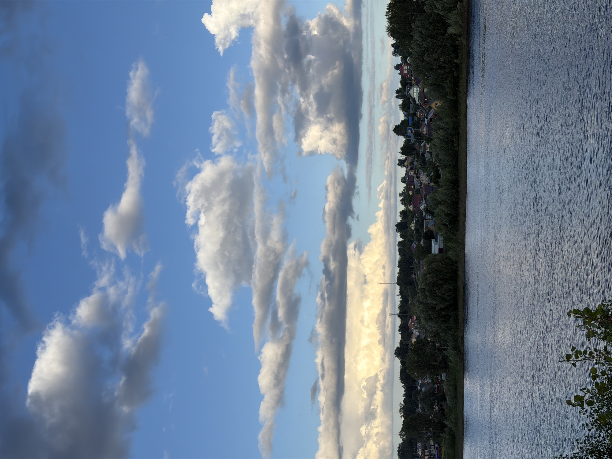 The image shows a wide body of water, likely a river, in the foreground. The water is a deep blue and has gentle ripples on its surface. Along the opposite bank, there is a densely wooded area with a village nestled within the trees. The village consists of numerous houses with red and brown roofs, interspersed with green trees. Above the village and trees, the sky is a bright blue, filled with a variety of fluffy white and gray clouds. The clouds are illuminated by sunlight, creating dramatic contrasts between light and shadow. In the far distance, on the horizon, a tall, slender tower, possibly a cell tower, is visible. The scene appears to be a tranquil, natural landscape with a human settlement integrated into it. This landscape is consistent with views of the Kama River in the city of Chaikovsky, Russia. There are no people or animals visible in the image.