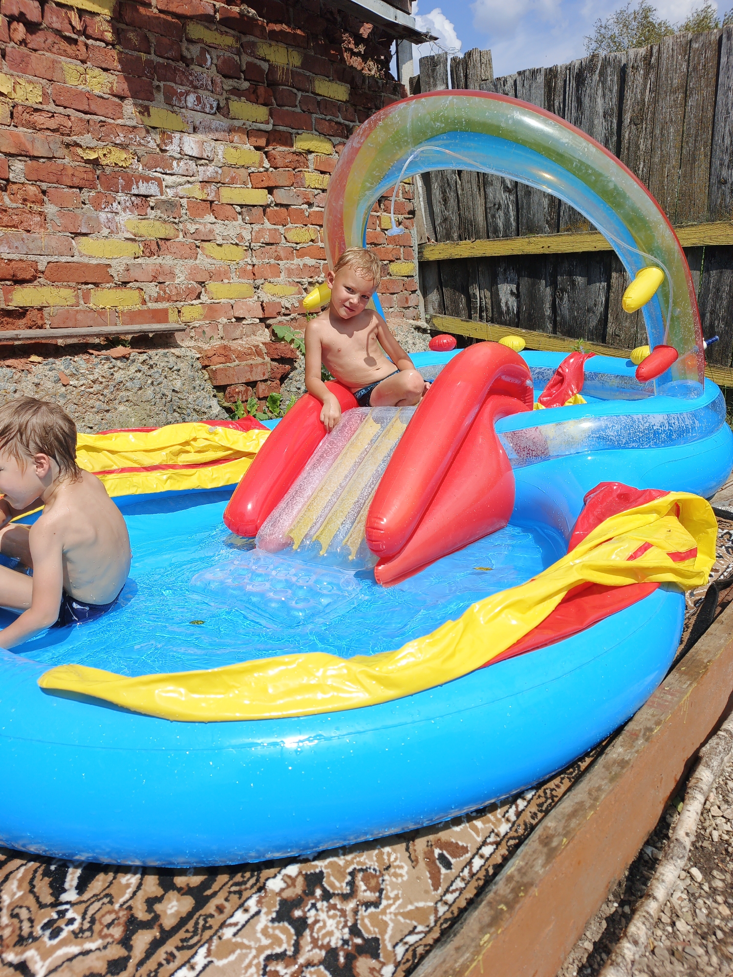 In an outdoor setting in Medvedyovka, Russia, two young boys are playing in a blue inflatable swimming pool. The pool features a red and yellow slide, a rainbow-shaped water spray arch, and yellow inflatable elements. One boy, with blond hair, is seated on the edge of the pool, while the other, also with blond hair, is on the slide. They are both shirtless and wearing swim trunks. The pool is situated on a patterned rug, with a rustic wooden fence and a brick wall in the background. The sky is partly cloudy.