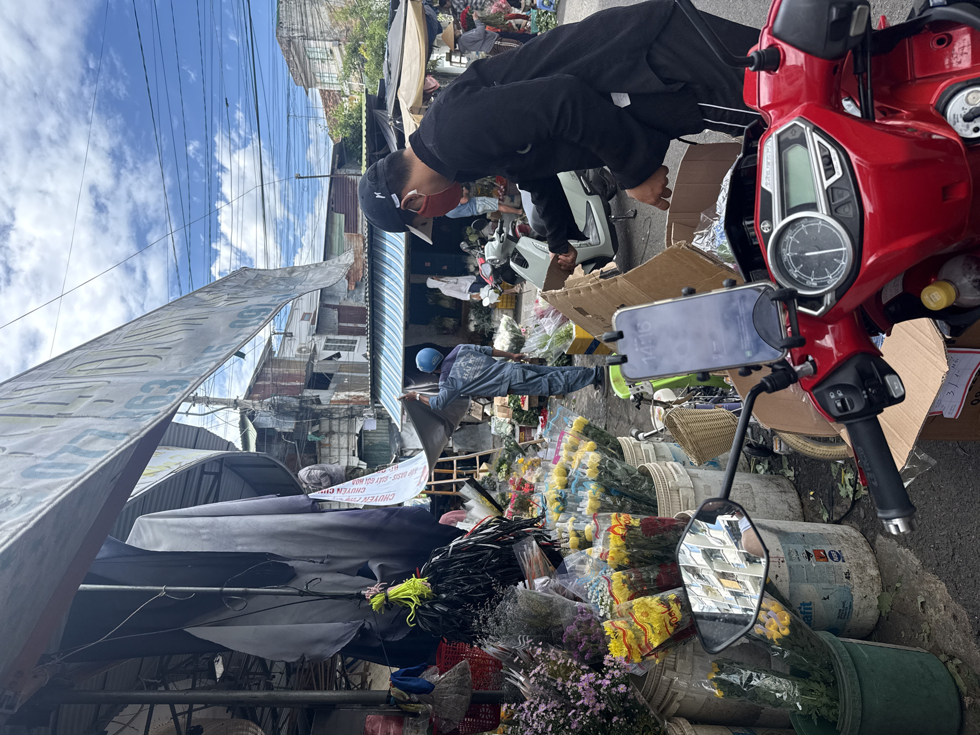 This image captures a bustling scene at a flower market, likely in Nha Trang, Vietnam. In the foreground, the handlebars and dashboard of a red motorcycle are visible, equipped with a phone holder displaying the time "14:16". A man wearing a black long-sleeved shirt, a baseball cap, and a red face mask is actively engaged in unpacking items from a cardboard box. He is positioned to the right of the motorcycle. Behind him, a variety of flowers are displayed in buckets and plastic packaging, including yellow roses and purple asters. Several other individuals are present in the background, one in a blue uniform appears to be setting up a tarp, and another person in a white hooded sweatshirt is near a scooter. The environment is outdoors, with a covered market stall structure overhead and buildings visible in the distance. The sky is partly cloudy, with power lines crisscrossing overhead. The overall impression is one of commerce and daily activity in an urban setting.