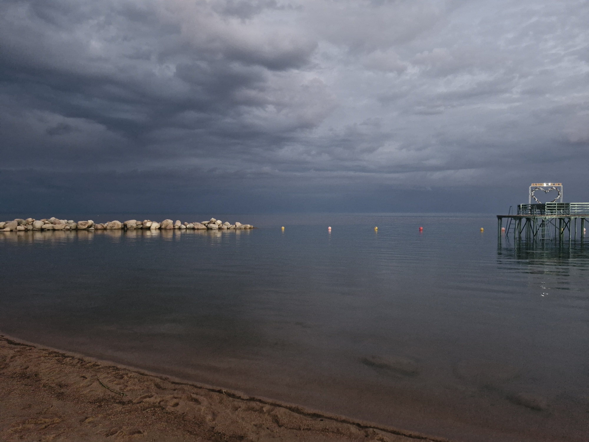The image depicts a cloudy, overcast sky over a tranquil body of water, likely a lake or sea, bordering a sandy beach. In the distance, a line of rocks forms a breakwater. To the right, a pier extends into the water, featuring a decorative heart-shaped structure at its end, possibly a wedding or proposal backdrop. Several buoys of red and yellow are scattered on the water's surface. The scene appears to be located near Bulan-Segettu, Kyrgyzstan, a region known for its natural beauty. No people are visible in the image.