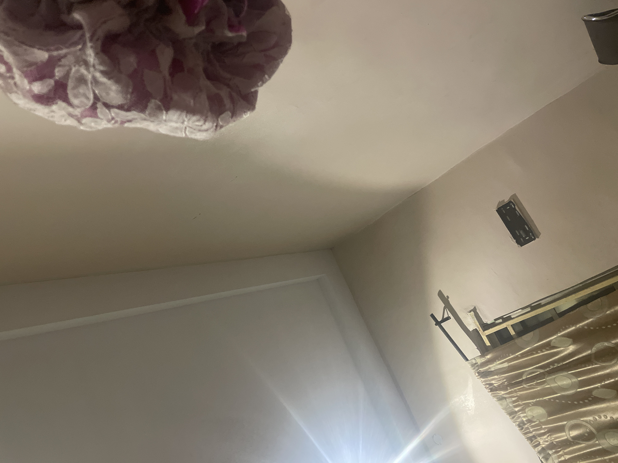 The image captures an indoor scene, likely a bedroom, from a low-angle perspective looking upwards. The ceiling is predominantly white, with a slightly recessed rectangular section. A bright light source, possibly a lamp or window, casts a lens flare across the upper left portion of the ceiling. On the right side of the frame, a decorative item, possibly a lampshade or fabric ornament with a floral pattern in pink and white, is suspended from above.
In the lower section of the image, a set of beige curtains with a circular pattern is drawn to the left. Behind the curtains, wooden beams are visible, suggesting they are part of a window frame or construction. A black metal curtain rod is mounted above the curtains. To the right of the curtains, mounted on the wall, is a black rectangular object, likely a soundbar or a media device bracket. The walls are painted a neutral, light beige color. The scene is dimly lit, with shadows cast by the curtain rod and the light source. The image was taken in Jalingo, Nigeria.