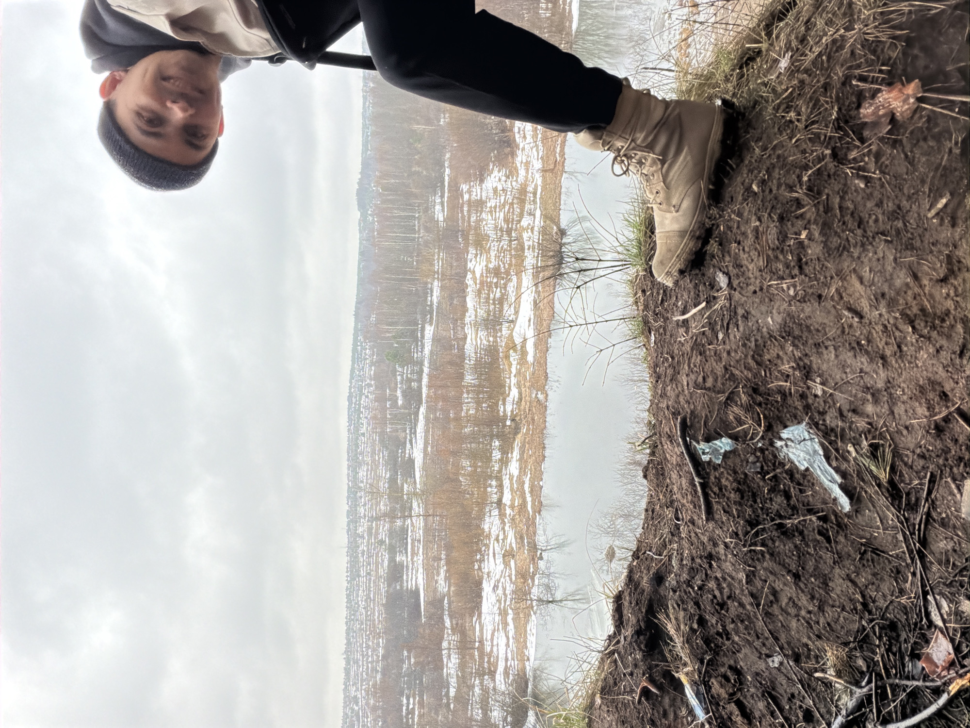 The image shows a person standing on the edge of a muddy bank overlooking a body of water, likely a river or a pond, in what appears to be a wooded area. The person, dressed in dark clothing, is wearing tan boots and a hat. Patches of snow cover the ground on the opposite bank. The sky is overcast. The location is near Izhevsk, Russia, based on the landscape and architectural features visible across the water. The ground is muddy with twigs, dead leaves, and litter visible. The image has been rotated 180 degrees.