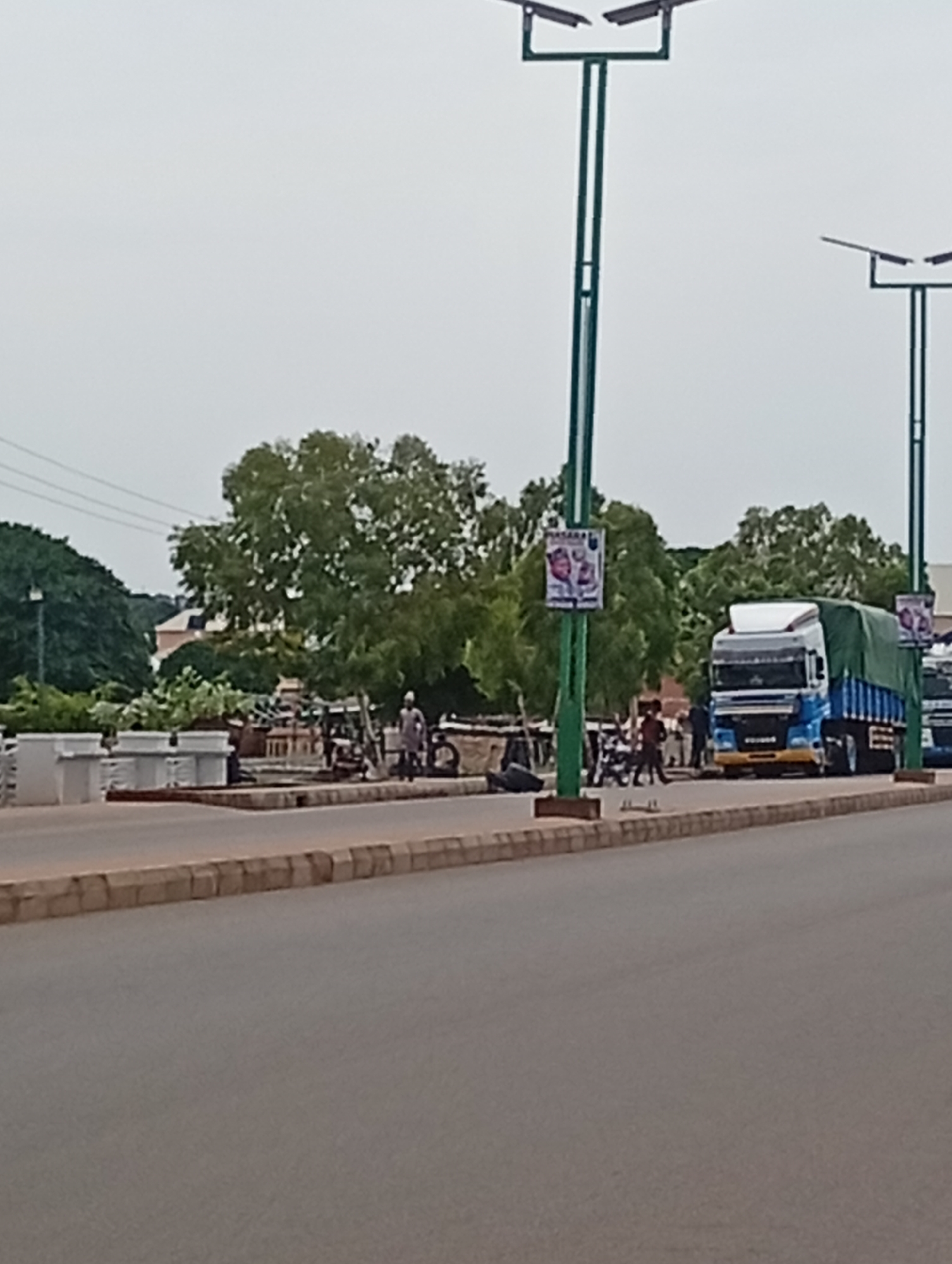 The image captures a street scene in Tumfure, Nigeria, under an overcast sky. Two tall, green lampposts with solar panels are prominently featured, one in the foreground to the left and another further back to the right. A large blue and white truck with a green tarp covering its cargo is parked on the right side of the road. Several people are visible on the sidewalk near the truck, some appearing to be interacting. Behind the lamppost on the left, a white concrete structure with planters is visible. Trees with lush green foliage are scattered throughout the background. The road in the foreground has a textured surface and is bordered by a concrete curb. Two posters are attached to the lampposts, one on each.