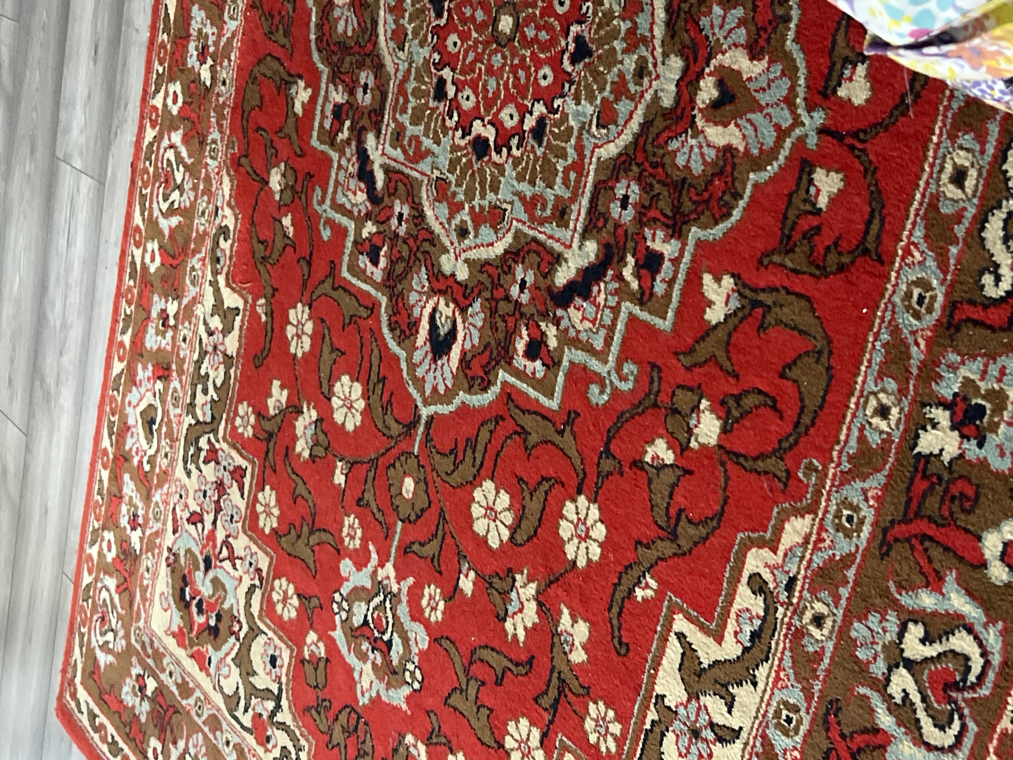 The image shows a close-up of a red oriental rug with intricate patterns. The patterns include floral designs in white and brown, as well as geometric shapes. The rug is likely indoors, and part of a light grey wooden floor is visible on the left edge. The location of the rug is unknown, and there are no people present.