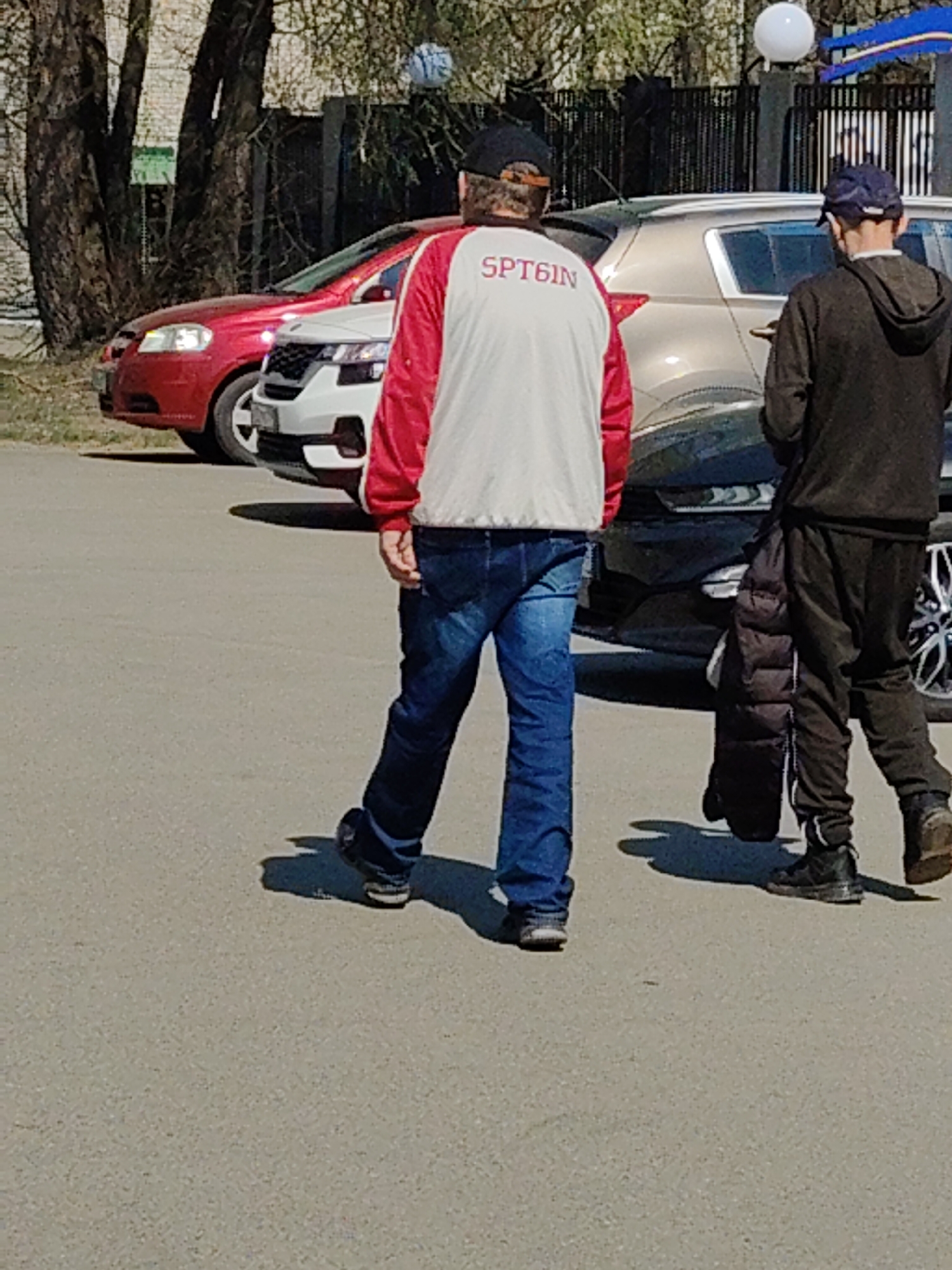 A medium shot depicts two adult males walking away from the viewer on an asphalt surface, with vehicles parked in the background, suggesting an urban setting in the city of Unknown, Unknown.
The individual on the left wears a baseball cap, a white jacket with red sleeves, blue jeans, and dark shoes. "SPTBIN" is printed on the back of his jacket. To the right, the second man dons a dark baseball cap, a brown hooded sweatshirt, dark sweatpants, and black boots. He carries a dark jacket in his left hand.
Parked behind the men are several vehicles: a red car, a white car, and a bronze or silver car. Trees are visible to the left, and a metal fence with a light fixture on top of it is on the right. The lighting suggests a sunny day. The composition is natural, focusing on the backs of the two individuals in motion, without any visible interaction between them.