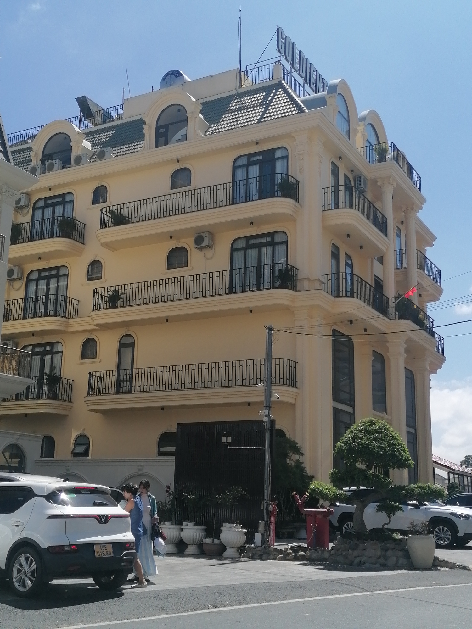This is a full-shot, eye-level, outdoor photograph of the Golden Hotel in Da Lat, Vietnam, taken on a sunny day. The hotel is a multi-story building with a light yellow facade, ornate architectural details, and numerous balconies with black railings. The name "GOLDEN" is prominently displayed in large, dark letters across the top of the building. The hotel has a distinctive green tiled roof with arched windows.
In the foreground, a white car with its trunk open is parked on the right side of the image. Two women are standing near the open trunk, engaged in conversation or activity. One woman is wearing a blue dress and the other is wearing a light blue jacket over a dress. Further to the right, a small, artfully pruned tree, possibly a bonsai, is situated on a bed of rocks. Several large, white planter pots are arranged in front of the building. A red fire hydrant and a red metal utility box are also visible on the left side of the image, next to a lamppost with a security camera. The background consists of a clear blue sky with a few scattered white clouds. The overall scene suggests a commercial establishment with accompanying outdoor elements.