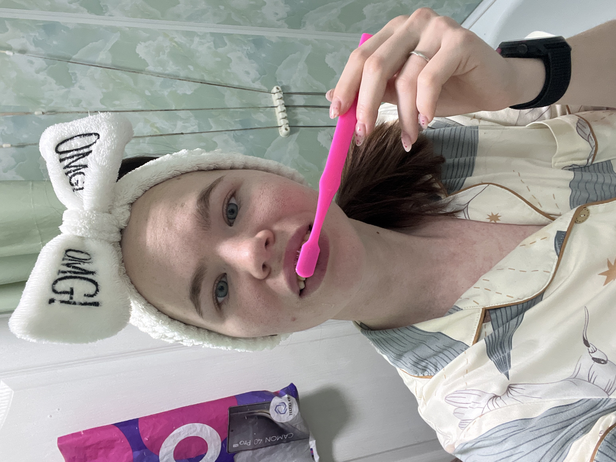A person is brushing their teeth in a bathroom. The person is wearing a white fluffy headband with "OMG!" written on it, and patterned pajamas. They are holding a pink toothbrush to their mouth. A black smartwatch is visible on their wrist. In the background are a white wall, green tiled wall, and a white fixture. A box for a Tecno Camon 40 Pro smartphone is in the foreground. The location is Chaikovsky, Russia.