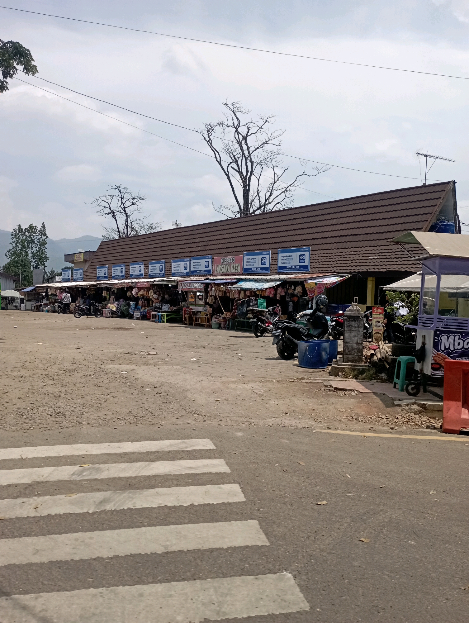 The image depicts a street scene in Panjalu, Indonesia, likely a market area. In the foreground, a pedestrian crossing is visible on an asphalt road. The midground is dominated by a long, single-story building with a dark brown, tiled roof, housing multiple small shops or stalls. These stalls are open to the street, displaying various goods, including food items and packaged products. Several signs are visible, with some featuring the "BRI" logo, indicating banking services. Motorcycles are parked in front of the stalls, suggesting transportation. A few individuals, dressed casually, are seen standing or walking among the stalls. The environment appears somewhat dusty, with loose gravel and debris on the ground. In the background, trees, some bare and others with foliage, frame the scene, and a mountainous landscape is faintly visible. The sky is overcast. To the right of the main building, a purple and white structure, possibly a food kiosk, is present with additional signage.