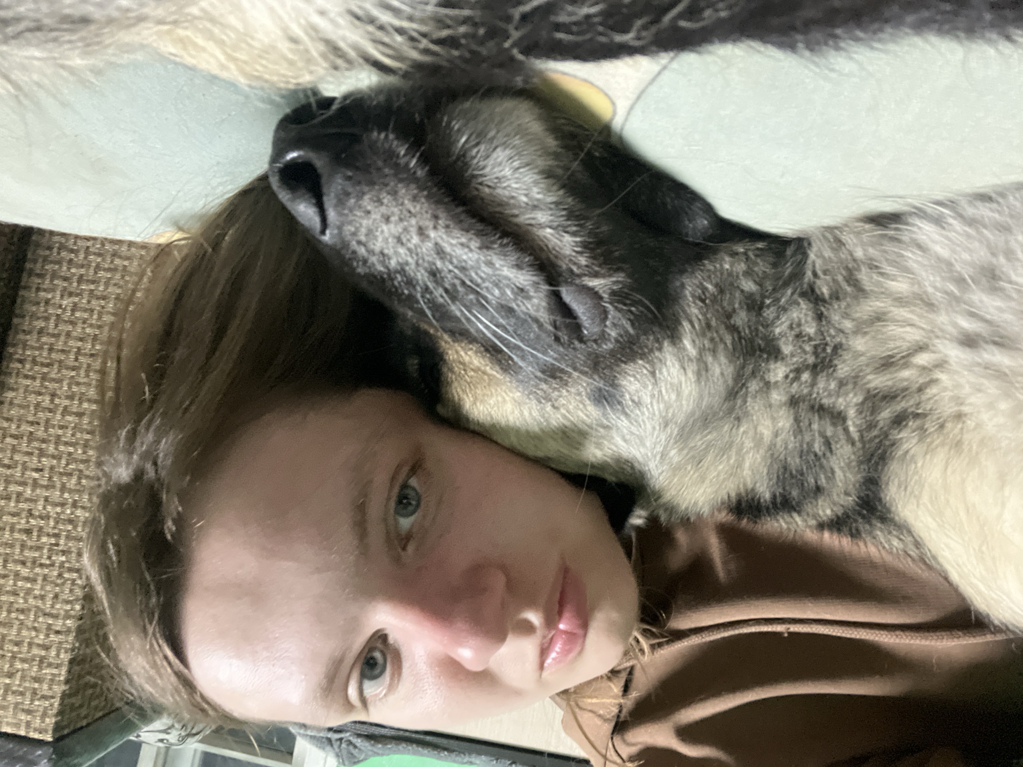 The image shows a close-up, rotated portrait of a young person and a dog, likely a selfie or a very close-up shot. The person, with fair skin, light-colored eyes, and brown hair, is lying on their side, looking directly at the camera with a neutral and relaxed expression. They are wearing a brown hooded top.
Resting closely beside the person's head and shoulder is a dog, possibly a husky or a similar breed, with a mix of gray, black, and white fur. The dog's head is visible, showing its dark nose and some whiskers, and it appears to be resting peacefully, with its eyes closed or partially closed.
The setting appears to be indoors, with a textured brown surface (possibly a sofa or chair armrest) visible on the left side of the frame and a light blue-green, soft surface (likely a pillow or blanket) behind the dog's head. The intimate framing suggests a moment of calm companionship between the person and their pet. No specific time of day or weather conditions are discernible from the indoor setting. There is no visible text in the image. The provided location context for this scene is Nizhny Tagil, Russia.