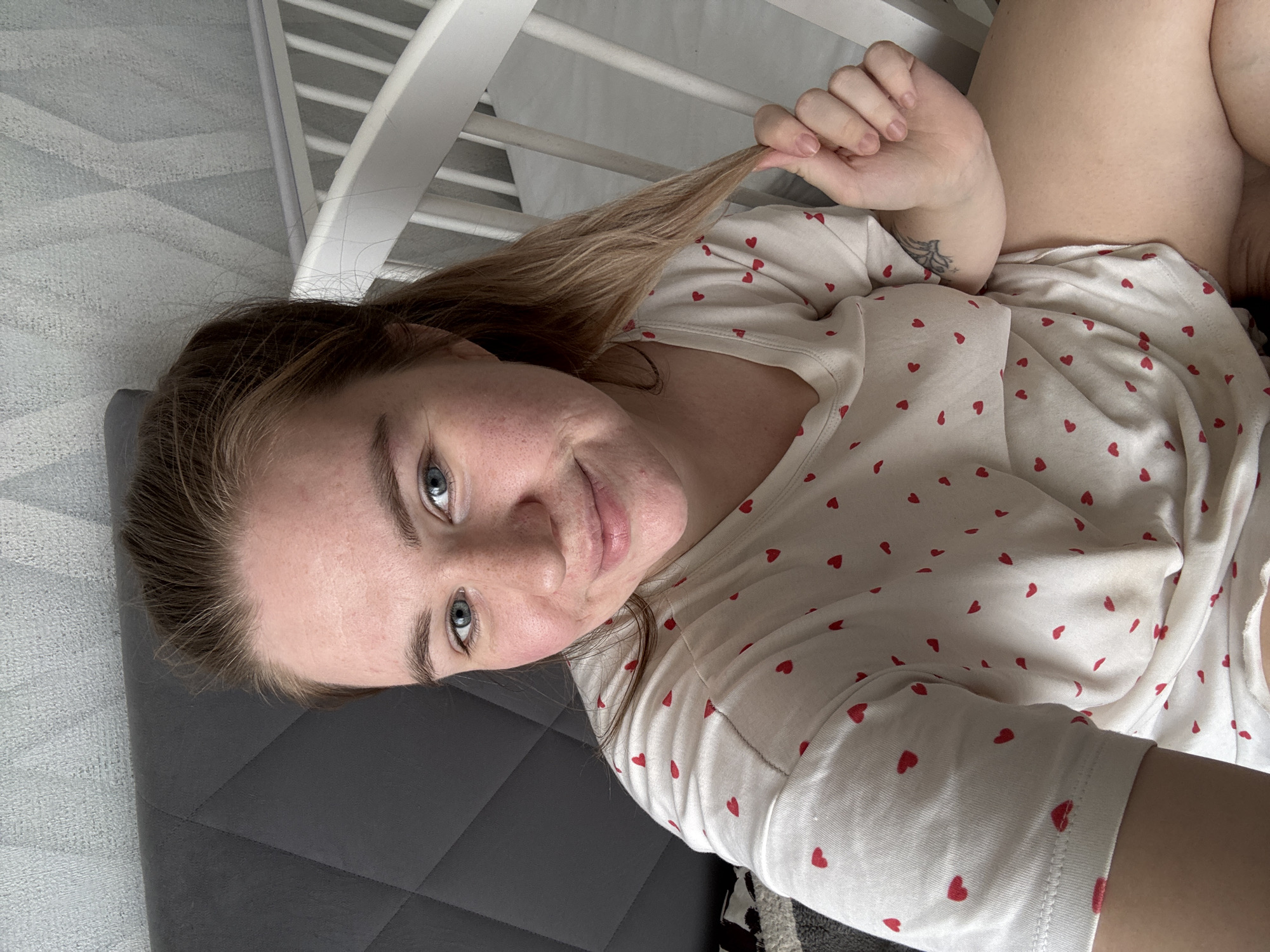 A woman with fair skin and blue eyes lies on a gray quilted surface. She is wearing a white t-shirt with red heart-shaped prints and holding a strand of her light brown hair. A white slatted structure, possibly a crib or railing, is partially visible in the background. The location is Chaikovsky, Russia.