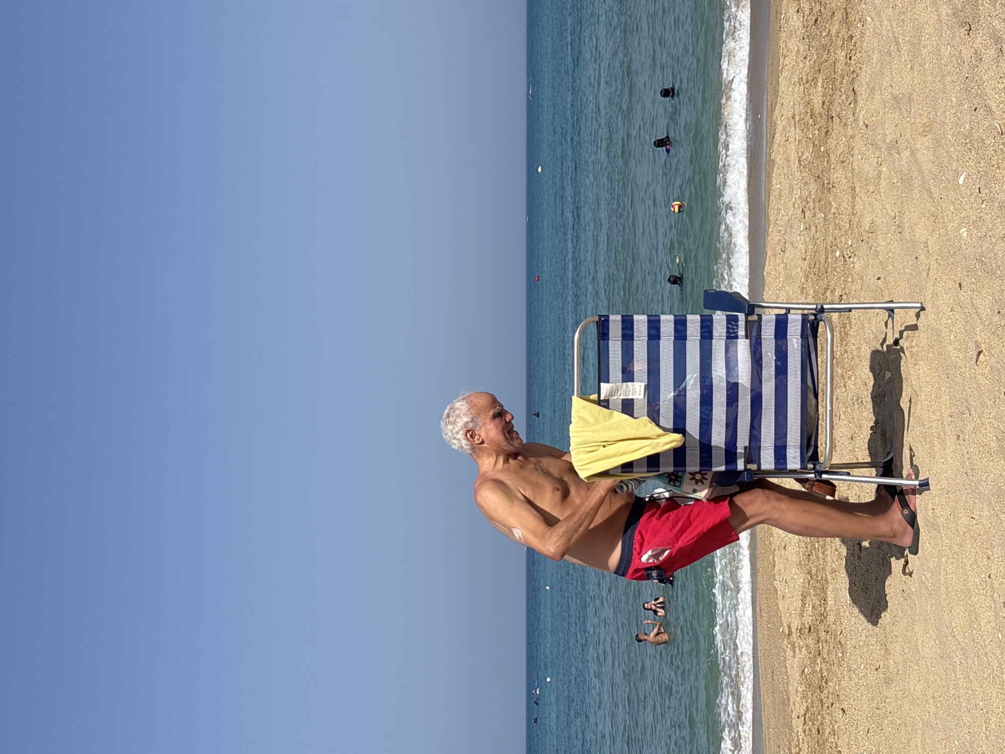 The image depicts a scene on a beach in M'Diq, Morocco. A man with gray hair and bare chest is sitting in a blue and white striped beach chair. He is wearing red swim trunks and black flip-flops. A yellow towel is draped over the armrest of his chair. He is looking to his left, and his expression suggests relaxation or contemplation.
The beach itself is sandy, with some shells visible. The ocean stretches out in front of the beach, with gentle waves breaking on the shore. Several people are visible in the water, some appearing to be swimming or wading. There are buoys in the water, suggesting a designated swimming area.
The sky above is clear and a vibrant blue, indicating a sunny day. The lighting suggests it is likely daytime, possibly late morning or afternoon. The overall atmosphere of the image is one of a peaceful and enjoyable day at the beach. There is no visible text in the image.