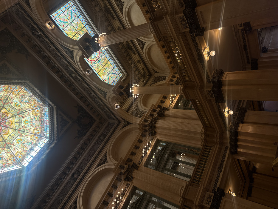 The image depicts the elaborate interior architecture of Galerías Pacífico, located at Florida 753 in Buenos Aires, Argentina. The scene is characterized by a grand, multi-tiered hall viewed from a low angle, emphasizing the ornate ceiling and upper levels.
A dominant feature is the large, octagonal stained-glass dome positioned centrally at the apex, allowing natural light to enter and creating prominent lens flares. Surrounding this dome, and extending throughout the upper structure, are richly decorated cornices, friezes, and archways adorned with intricate relief work in dark and light tones. Several smaller, arched stained-glass windows, featuring geometric and floral patterns in vibrant colors, are visible on the upper levels.
The structure is composed of light-colored, possibly marble or stone, walls and columns with fluted shafts and ornate capitals. Multiple balconies with dark, decorative balustrades span the upper floors. Clusters of spherical light bulbs, forming part of decorative fixtures, illuminate various sections of the architecture, casting bright glows and additional lens flares. Large, dark-framed window-like openings with interior reflections are present on the visible lower levels. No individuals are present in the depicted environment.