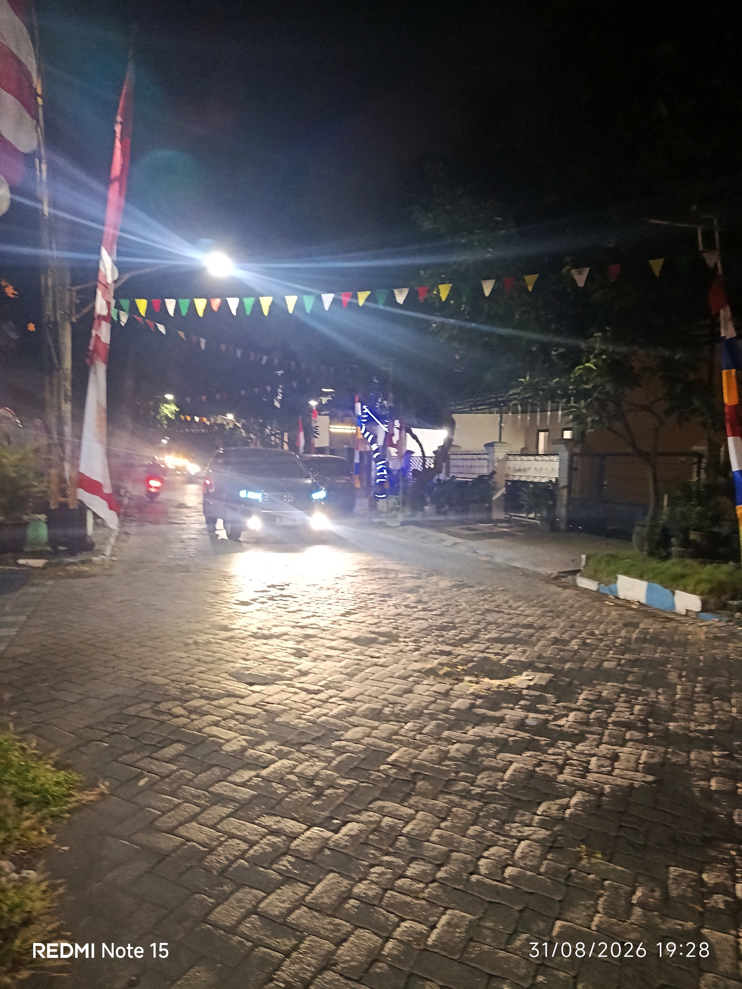 The image shows a street scene at night in Keboansikep, Indonesia. A dark-colored car with its headlights on is approaching the viewer on a cobblestone street. In the background, there are buildings decorated with colorful triangular flags and lights. To the left, a white and red flag is visible. The scene appears to be set during an event or celebration. The timestamp at the bottom right indicates the photo was taken on August 31, 2026, at 19:28. The bottom left corner shows "REDMI Note 15".