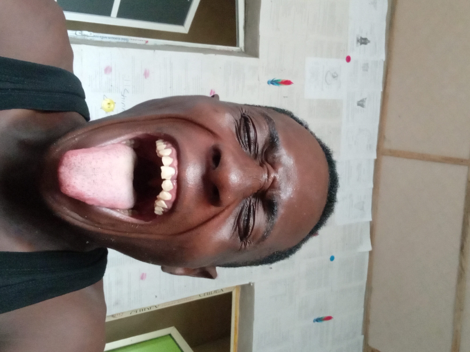 A close-up portrait of a dark-skinned person with their mouth wide open and tongue extended, eyes closed. The person is wearing a black tank top. The background features a wall covered in paper, with various drawings and colorful dots, and a window frame is partially visible. A wooden structure is also noticeable on the right side of the frame.
The location is unspecified but is reportedly in the city of Akwete, Nigeria.