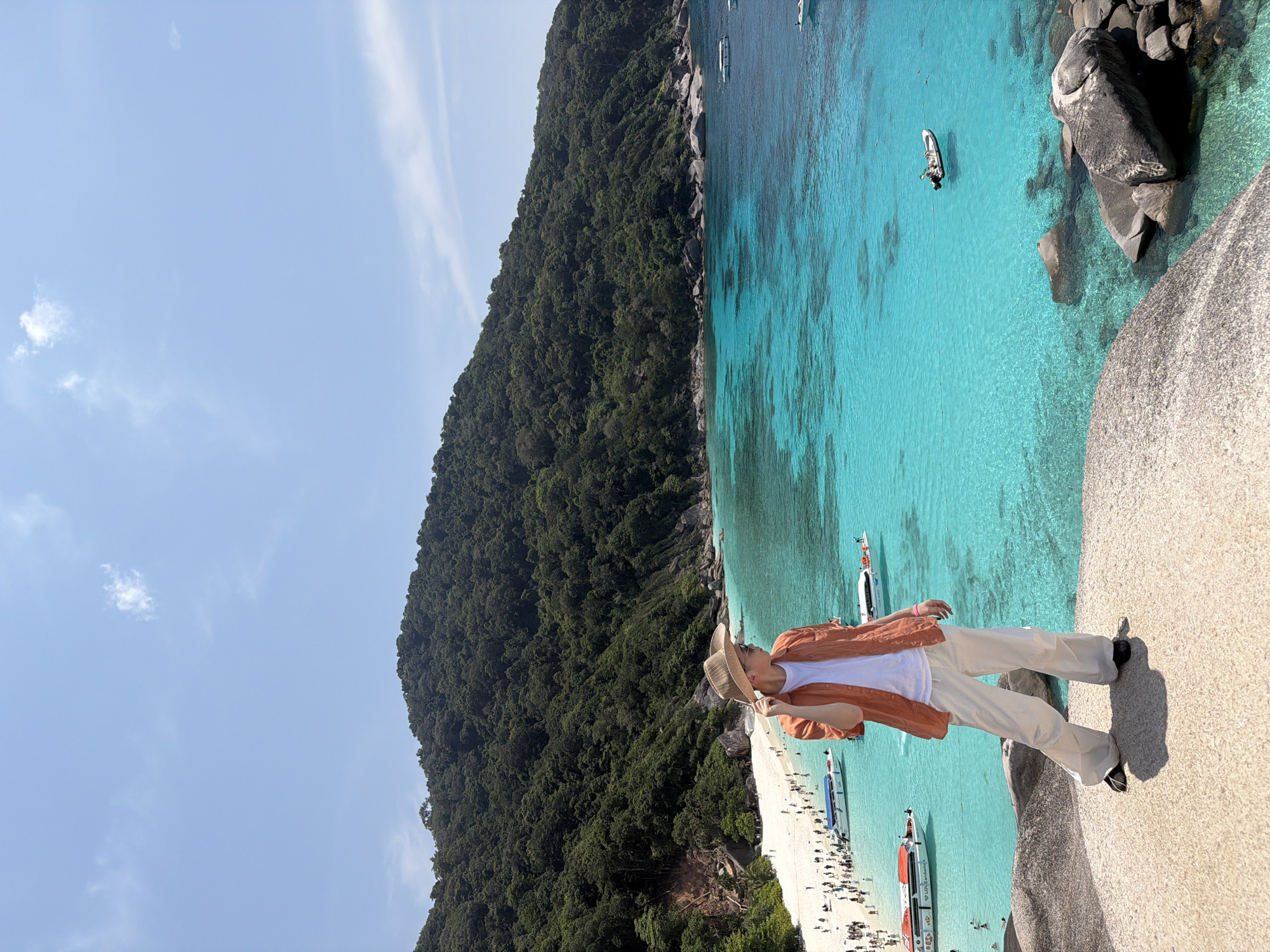 This image depicts a sunny tropical island landscape, identified as one of the Similan Islands, accessible from the coast near Lam Kaen, Thailand. In the foreground, a single individual stands on a large, light-colored granite rock formation. This person, appearing male, is wearing a white t-shirt, an unbuttoned orange short-sleeved collared shirt, light-colored wide-leg pants, a straw-style wide-brimmed hat, and sunglasses. The individual is positioned facing right, with their right hand touching the brim of the hat.
To the left of the foreground subject, a crescent-shaped white sand beach is heavily populated with numerous individuals. Multiple speedboats are beached or anchored near the shore, including vessels bearing the labels "LOVE Andaman" and "Sea Star". The water immediately adjacent to the beach is clear and bright turquoise, gradually transitioning to a deeper blue further from the shore. Many people are visible wading or swimming in the shallow waters near the beach. Further out in the bay, several other boats, including a small inflatable tender with occupants, are anchored.
The background consists of a prominent, steep hill densely covered in dark green tropical vegetation. The sky above is clear and light blue, with a few minimal, scattered white clouds. Large, dark granite boulders are present in the clear water and along the rocky coastline to the right of the foreground subject. The scene is illuminated by bright, direct sunlight.