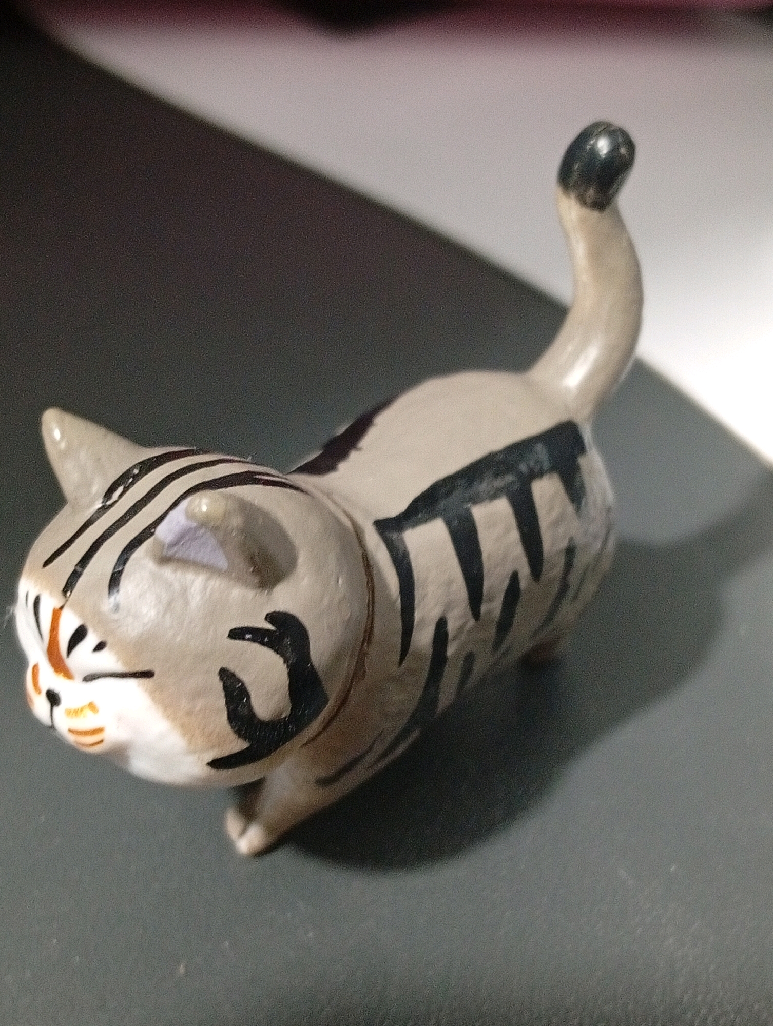 An overhead, slightly angled close-up shot shows a small, stylized ceramic cat figurine. The cat is a light tan color with black stripes painted on its back, head, and tail. It has a white chest and stomach area, with orange accents on its cheeks and nose. The cat's tail is curved upwards and ends in a black tip. The figurine is positioned on a dark gray surface, with a blurred, lighter surface and a dark shadow behind it. The lighting suggests an indoor environment. The location is not specified in the image, so the city is Unknown, Unknown.