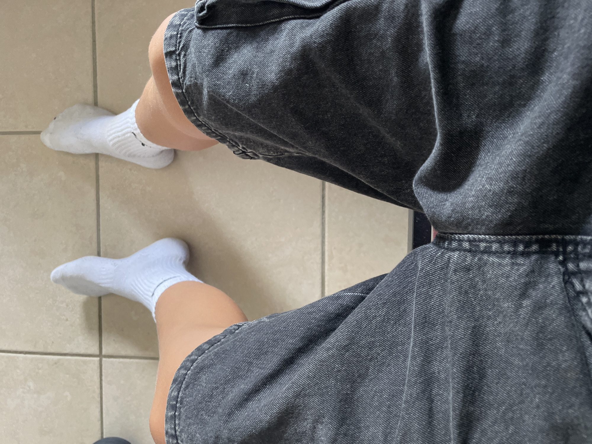 The image shows a first-person perspective of a person's legs wearing white athletic socks and dark wash denim shorts. The person is sitting down on a tiled floor. The lighting is bright, suggesting an indoor or outdoor setting during the day. The tiles are square and light-colored with dark grout lines. The denim shorts are loose-fitting and appear to be faded in places. The white socks have ribbed cuffs and a small black logo visible on the left sock. The person's skin tone is light.