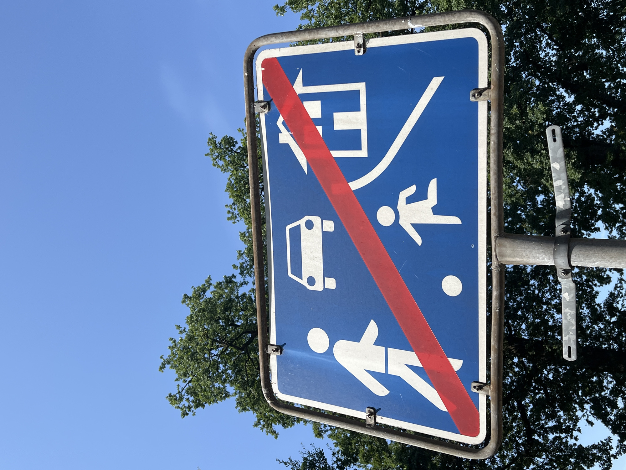 An outdoor, low-angle, medium shot captures a blue square sign mounted on a metal pole against a bright blue sky and a backdrop of lush green trees. The sign features white pictograms: a person walking, a car, a house, and a child running. A diagonal red stripe cuts across the sign, indicating the end of a restriction. The sign has a rounded metal frame. The pole has a metal bracket attached. The overall impression is a clear, sunny day.